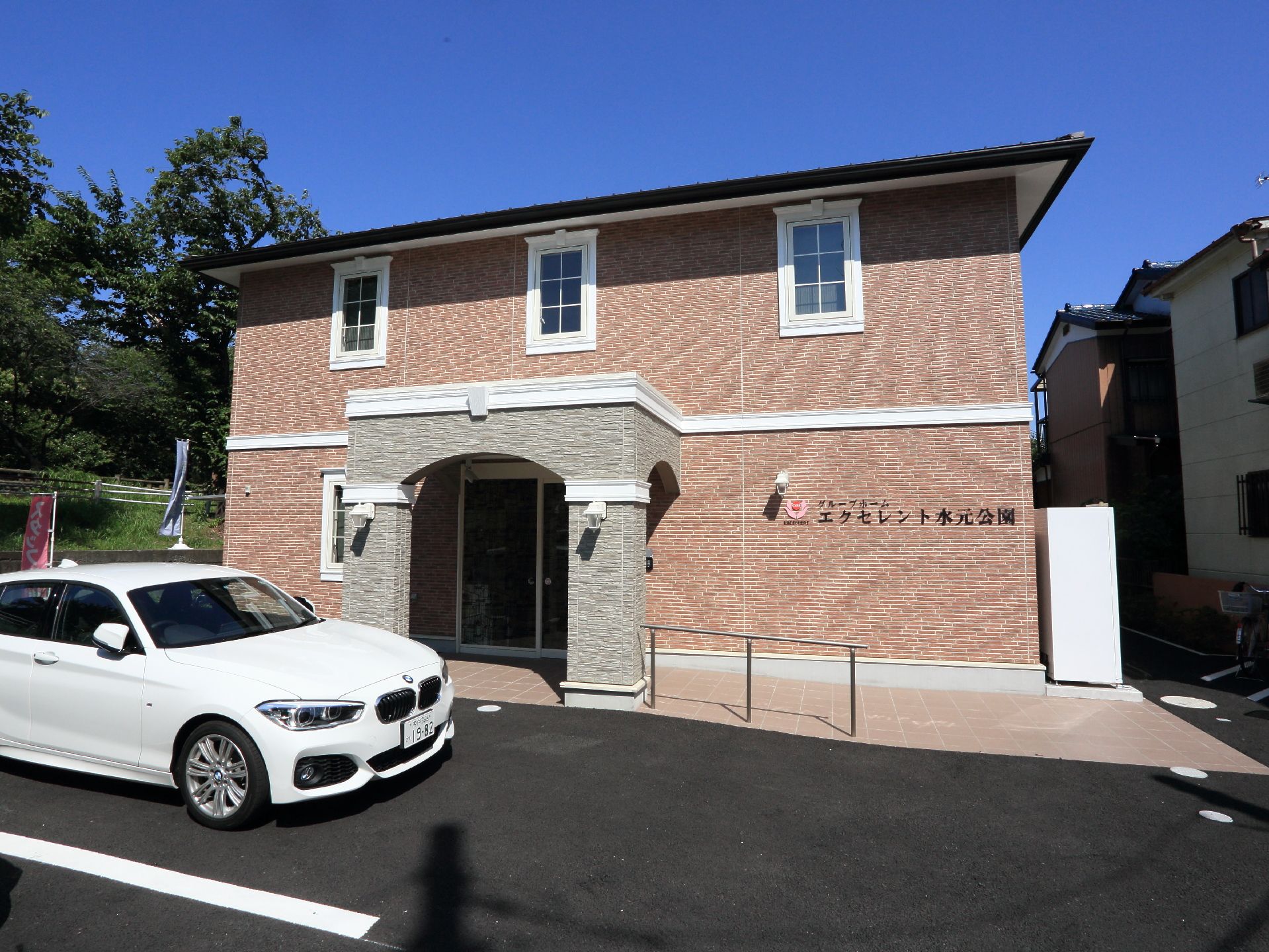施設のわきには植栽がしてあり、庭のようになっている。木造2階建ての建築物で茶色の外壁に白い窓枠である。