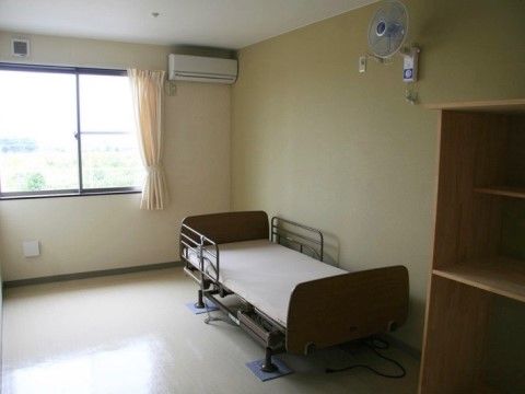 やや薄暗い室内の右手前に収納棚があり、その奥に介護用ベッドが置かれている。ベッドの上には壁掛け式の扇風機が取り付けられている。