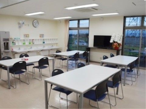 テーブルが間隔を開けて並んでおり、それぞれに会議用椅子が４脚ずつ置かれている。左奥には給茶機と手洗いが並んでいる。