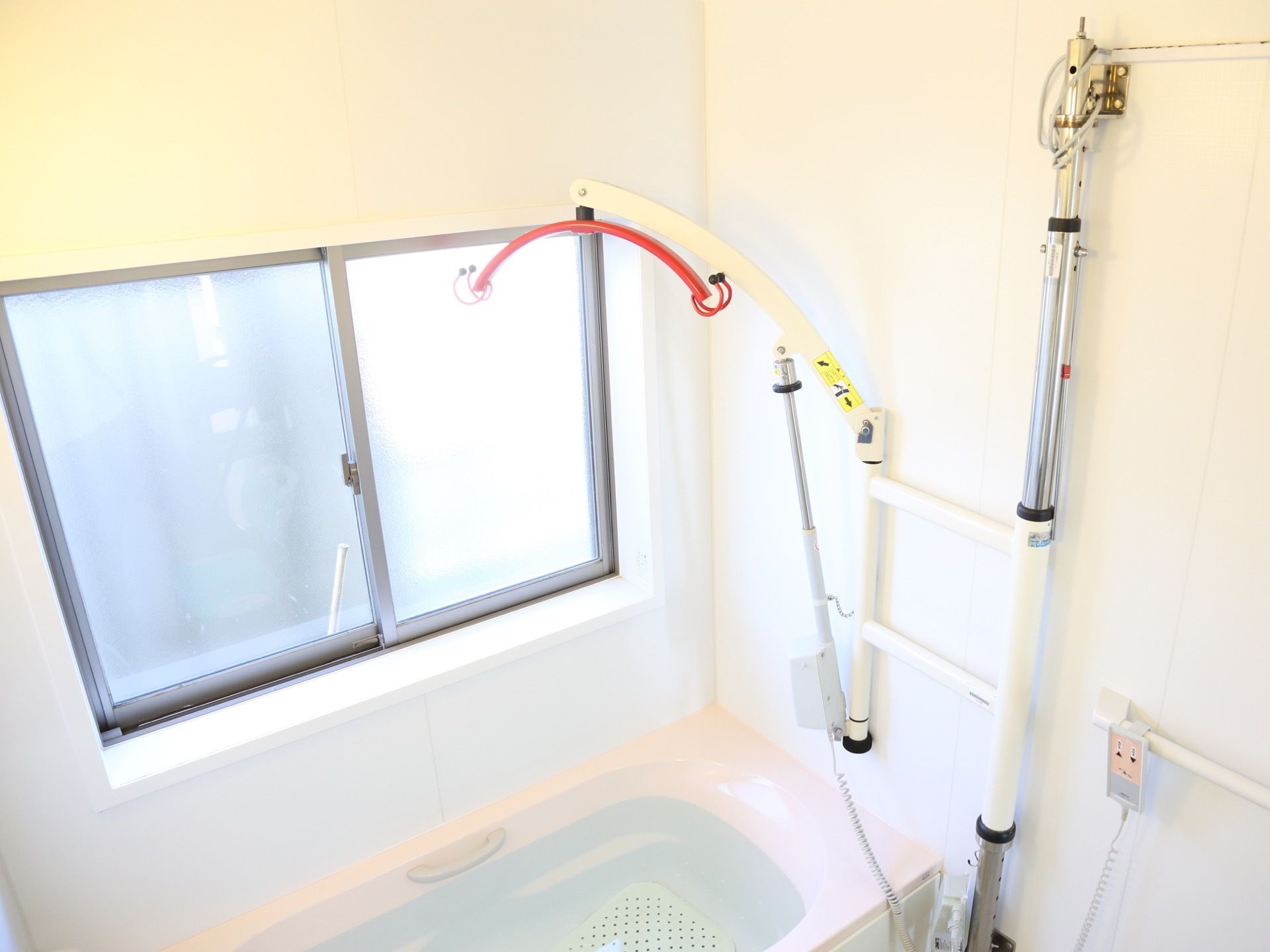 浴室の壁に入浴を介助する機械が設置されている。リフトを動かすリモコンが手すりに付けられている。浴室には大きな窓がある。