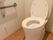 トイレスペースは白い壁とブラウンの床。便座に座って右側には手すりがあり、そのすぐ下には呼び出し用の緑色のボタンがあります。