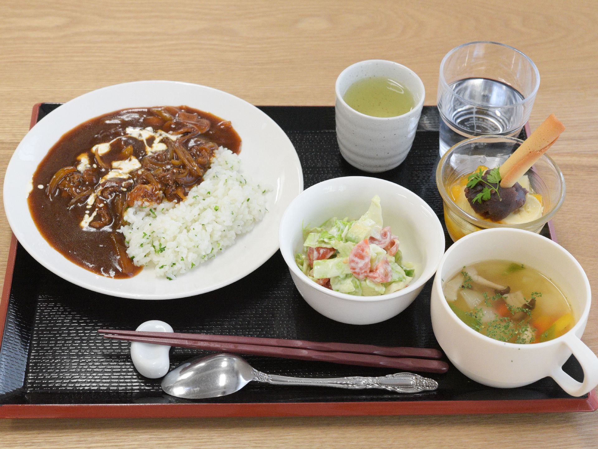 ご飯とカレーに一工夫のある主食にコールスローサラダ、スープ、デザート、水とお茶がお盆に乗せられている。