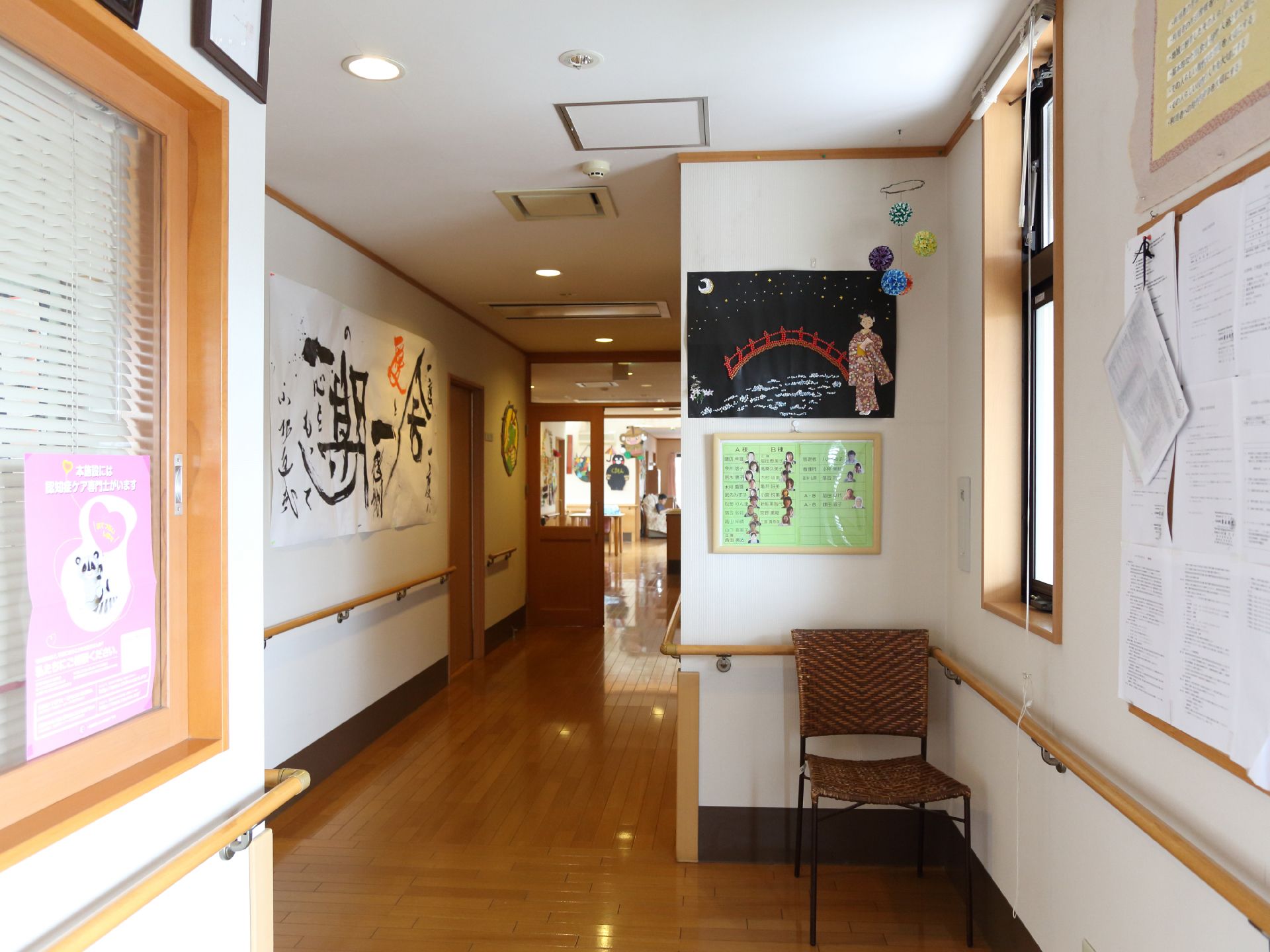 壁面にはたくさんの掲示物が貼付されている。手すりが設置されている。イスが1脚置いてある。手前の窓口にはブラインドが下りている。