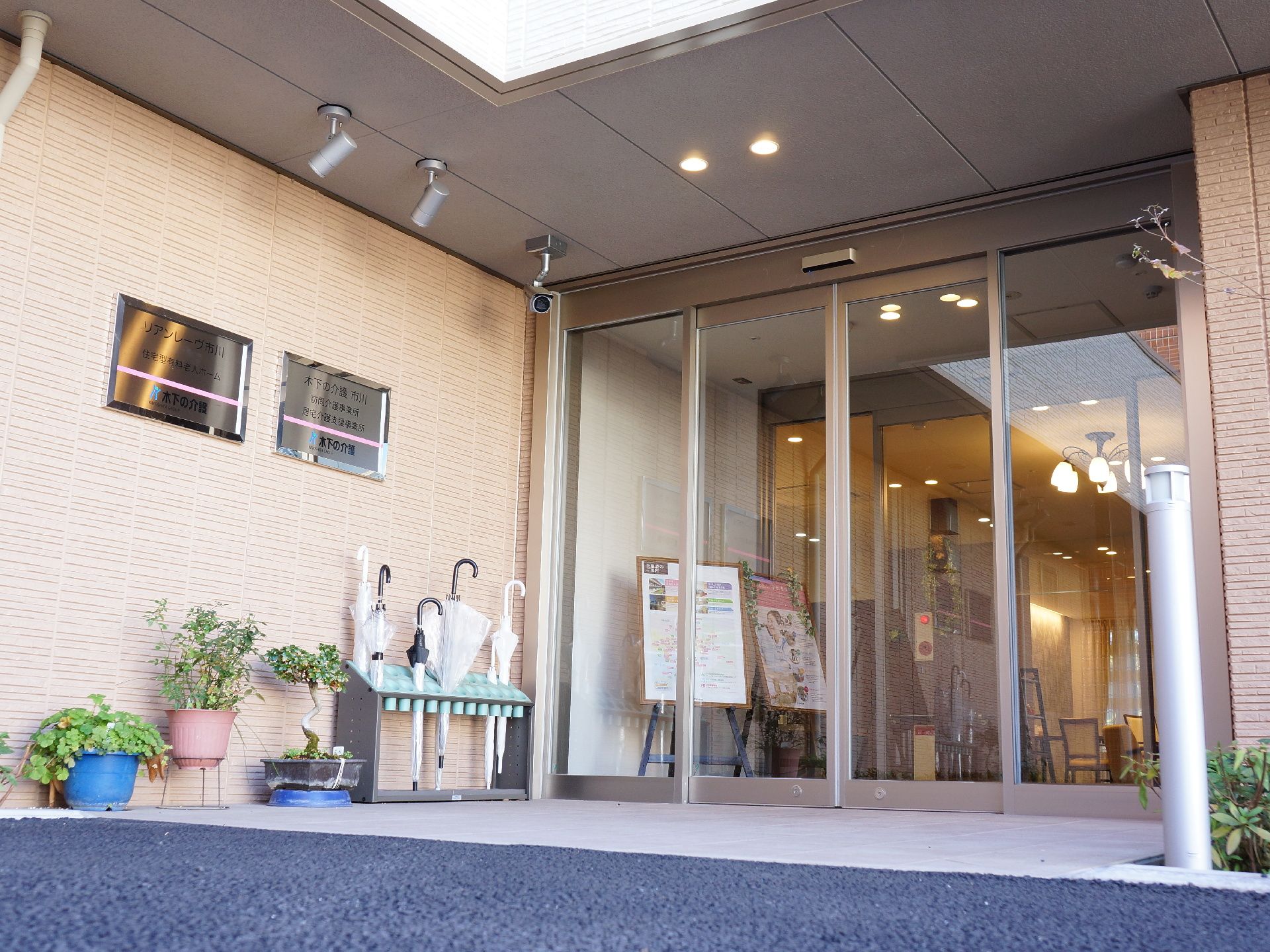 全面ガラス張りの自動ドアの玄関である。奥にもう一つ自動ドアがある。入り口横には傘立てや盆栽などが置いてある。防犯カメラもついている。