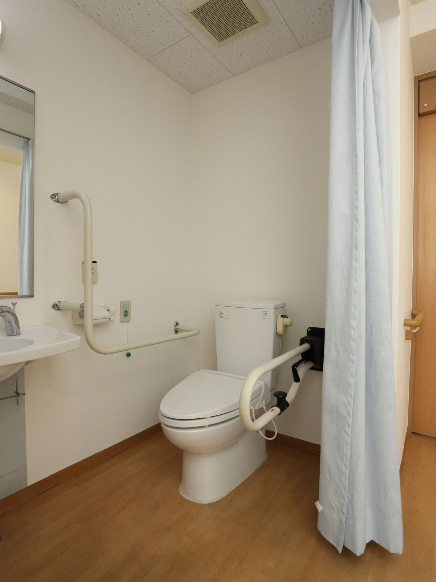 トイレはカーテンで仕切られている。壁面に手すり、緊急ボタンが設置されて安全面にも配慮されている。広い造りである。