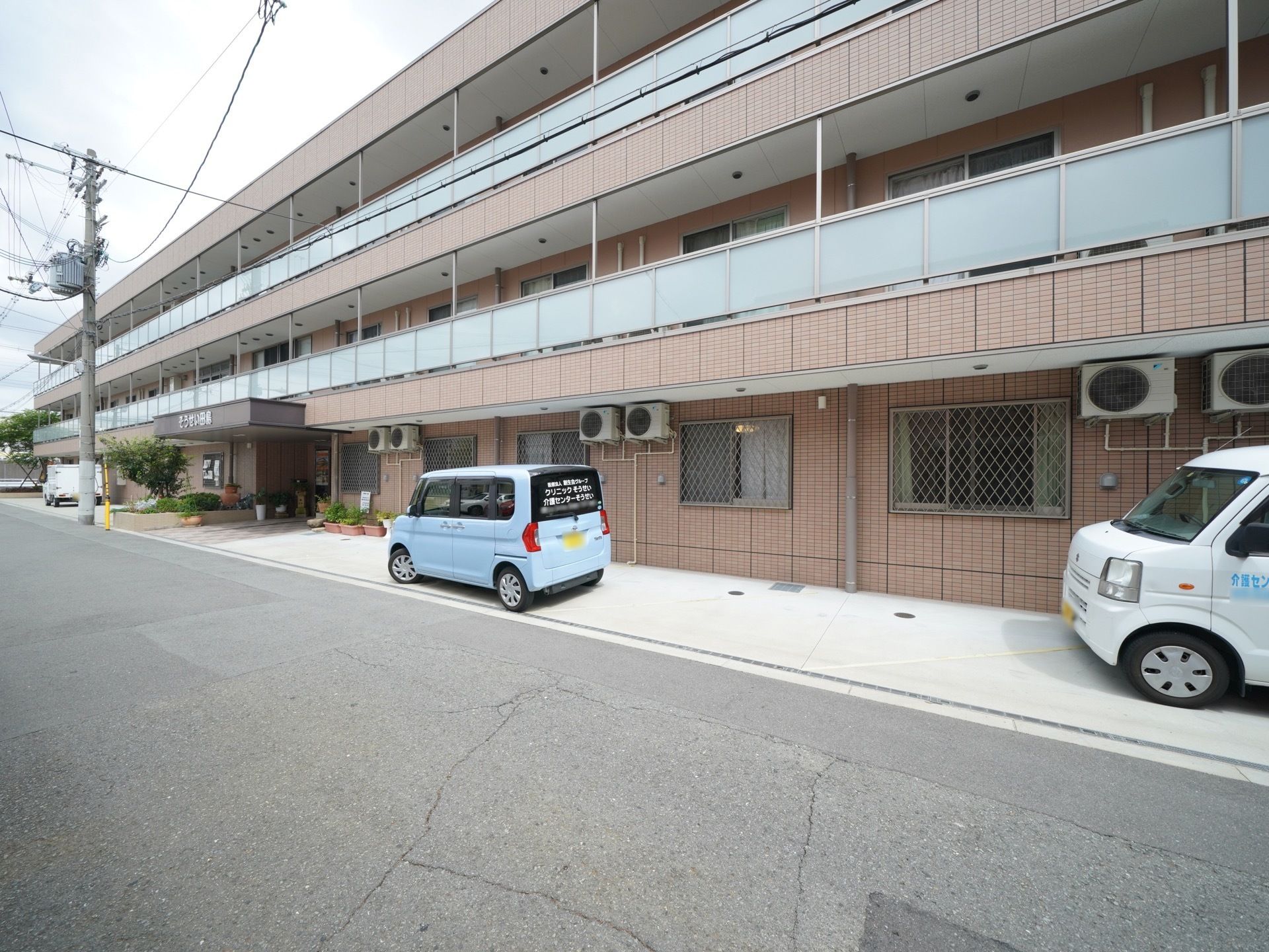 建物のすぐ目の前は道路がはしっている。敷地内に軽四自動車が２台、縦列駐車している。段差はなくフラットである。