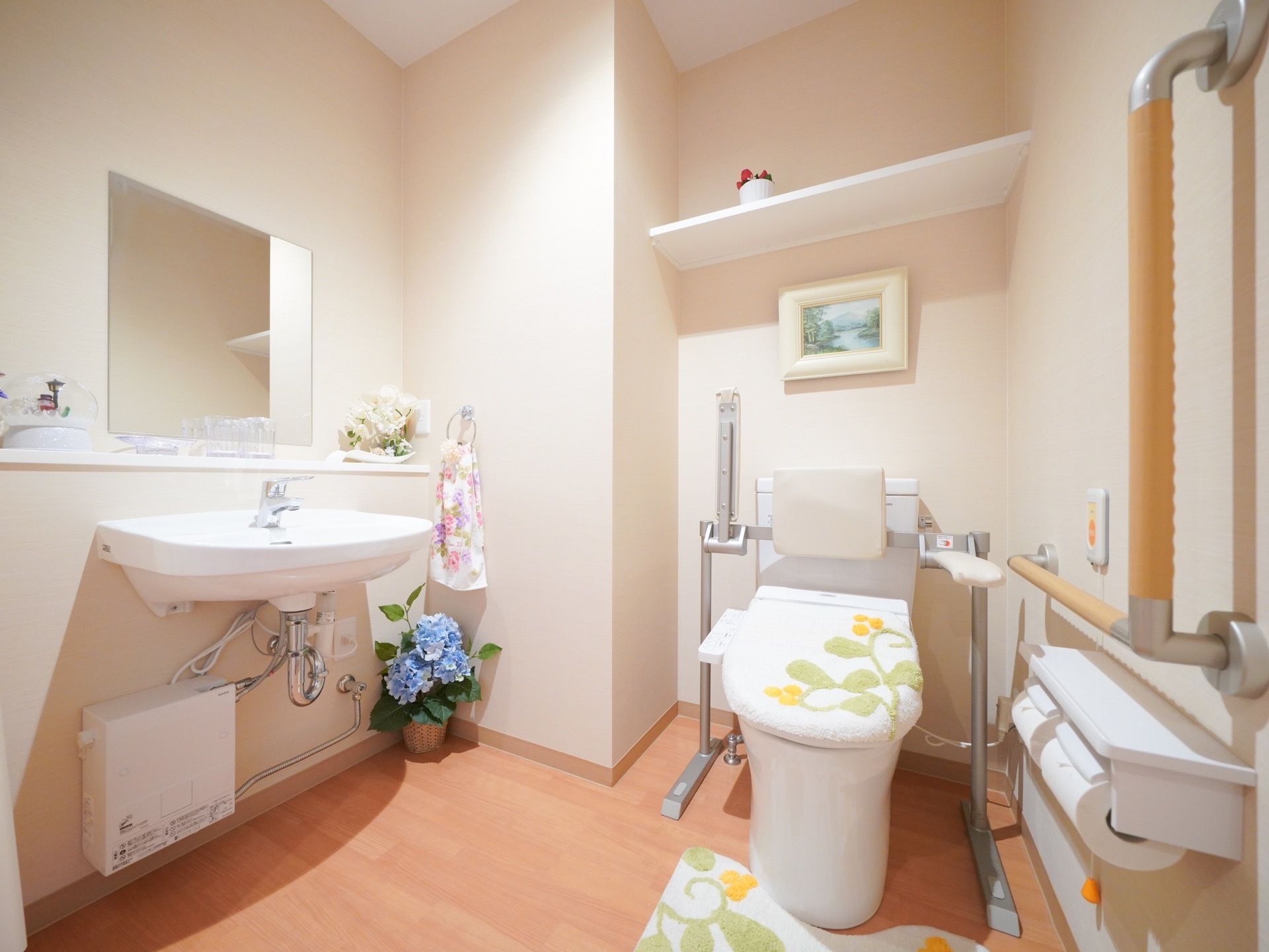 洋式便座に植物模様のカバー、マットがセットされている。手洗い場が設置されているほか、手すりも完備している。