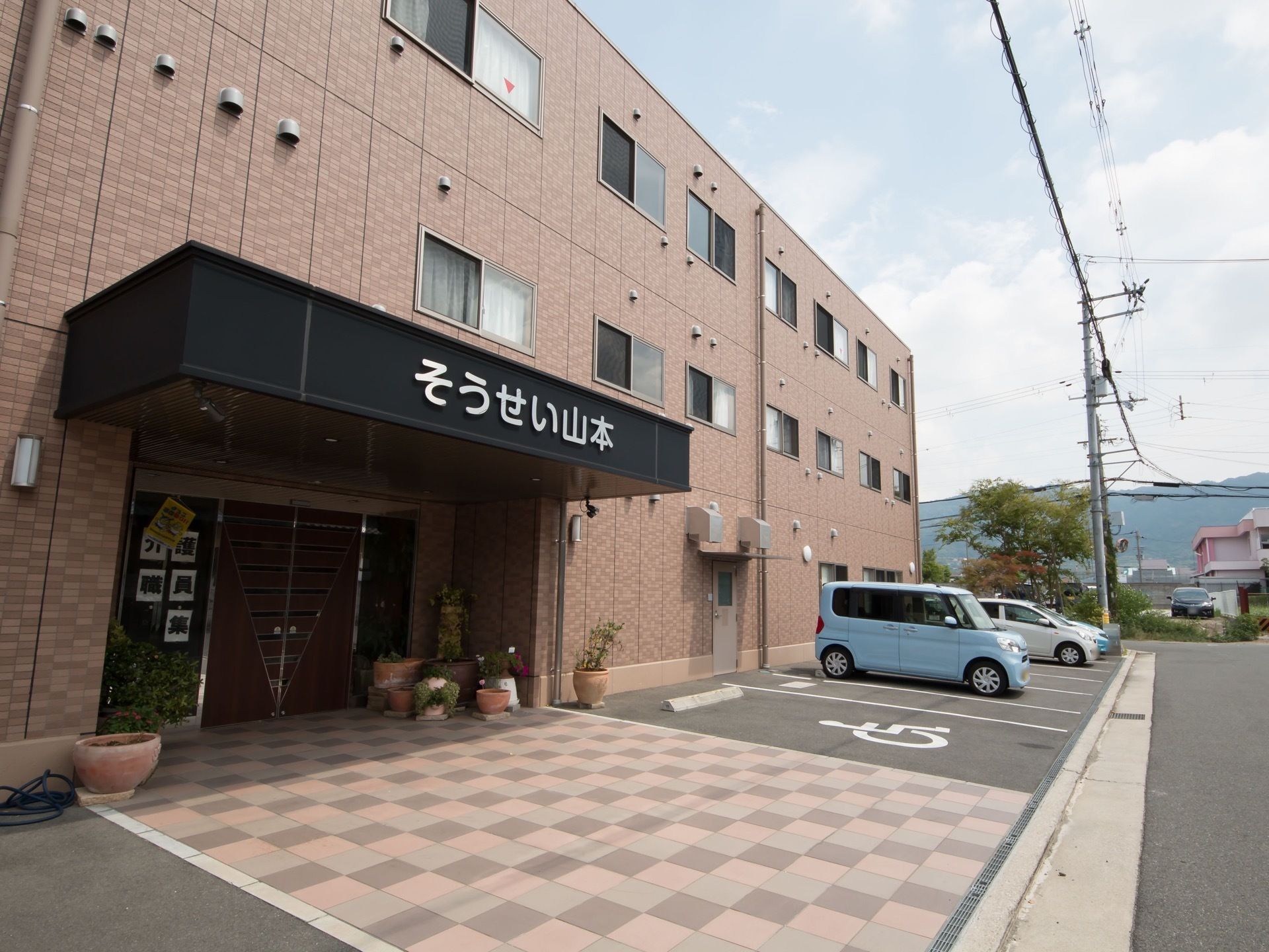 3階建ての施設の建物は、茶系のレンガを基調とした外観である。玄関の自動ドアの前には大きな屋根が備わったスペースがある。