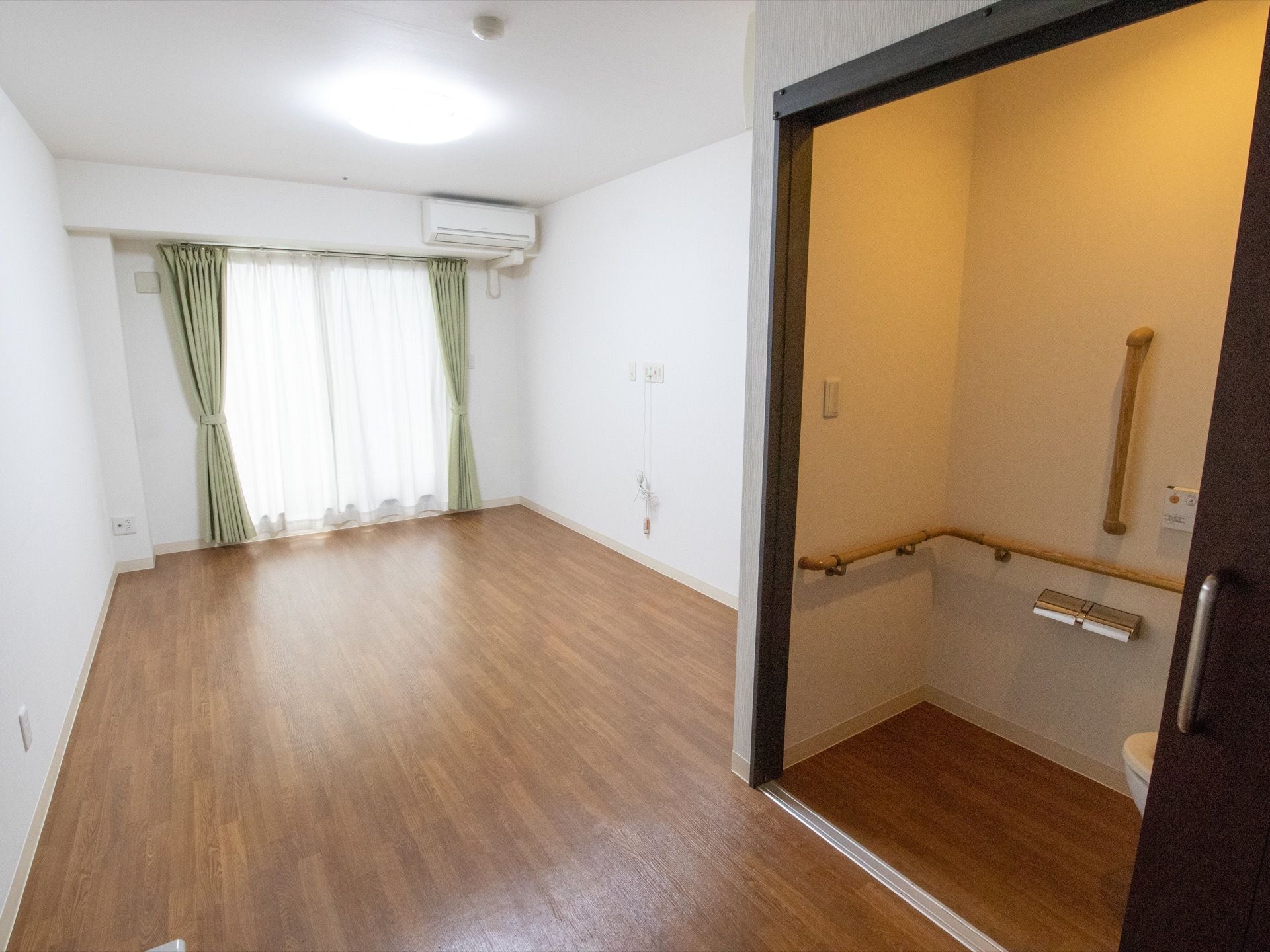 木目調の床に白い壁、グリーンのカーテンで落ち着いた雰囲気の居室である。室内にはトイレも設置されている。