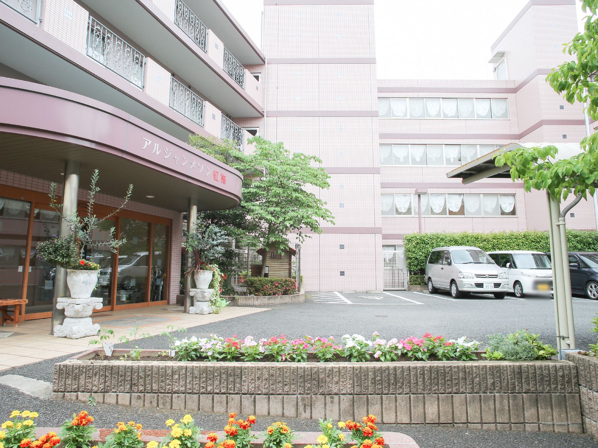 施設の建物は4階建てで、淡いピンクを基調としたデザインとなっている。玄関前には駐車スペースがあり、花が植えられている。
