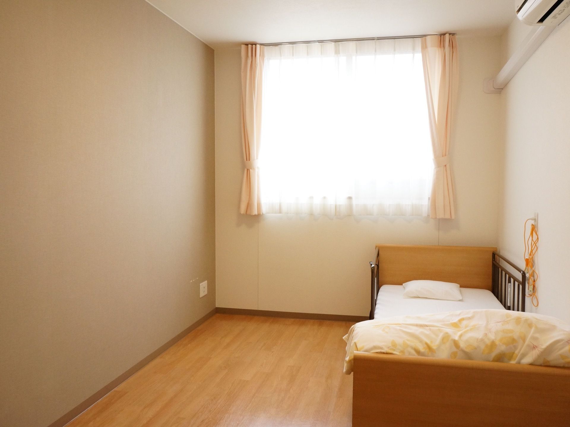 大きな窓がある居室は、光が射し込む明るい空間である。壁際に介護用ベッドを備え、横の壁にナースコールを設置している。