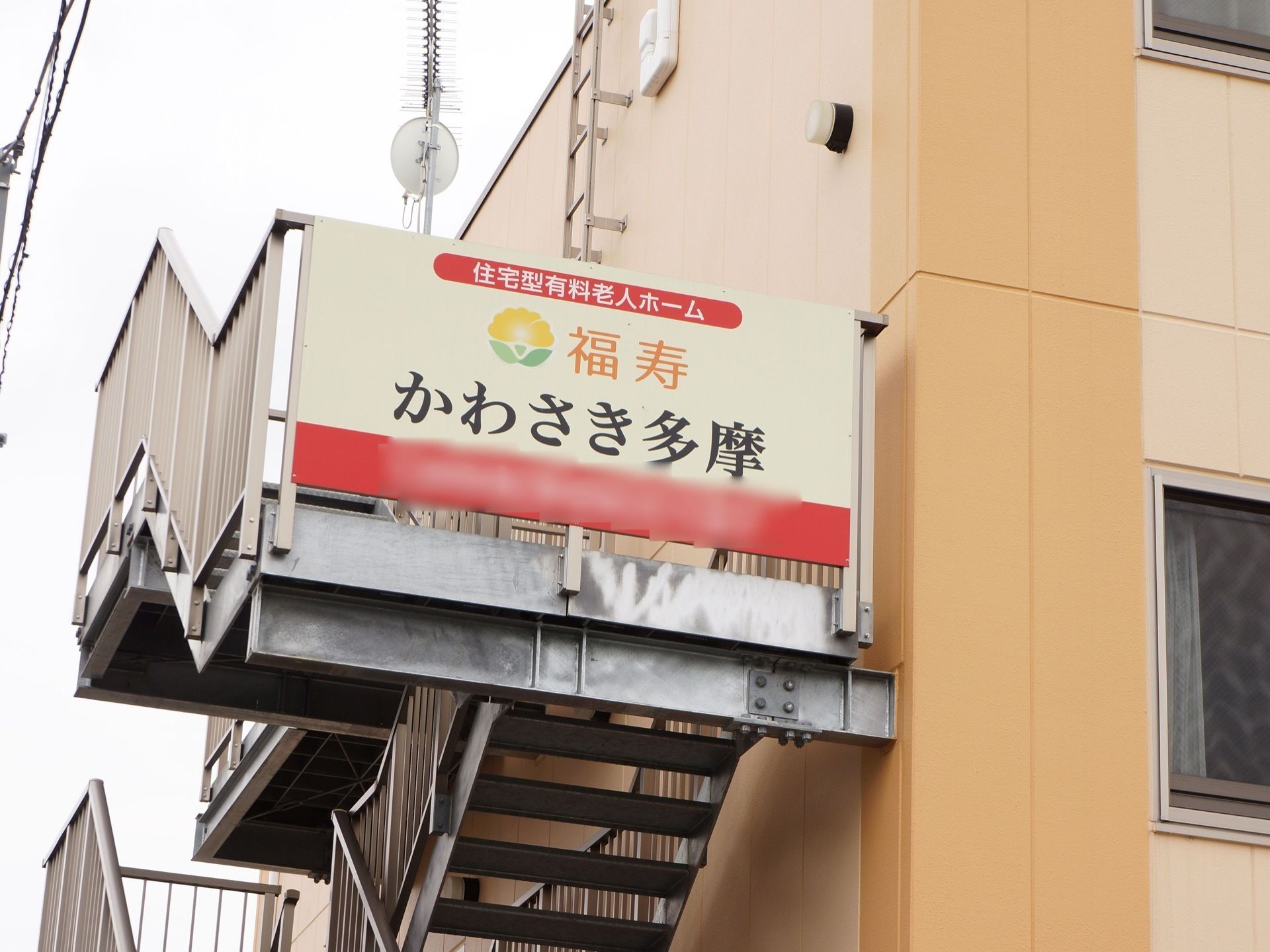 建物の横側に設置した階段のフェンスに大きな看板を掲げている。クリーム色の背景にオレンジと黒の文字の施設名とロゴを表示している。