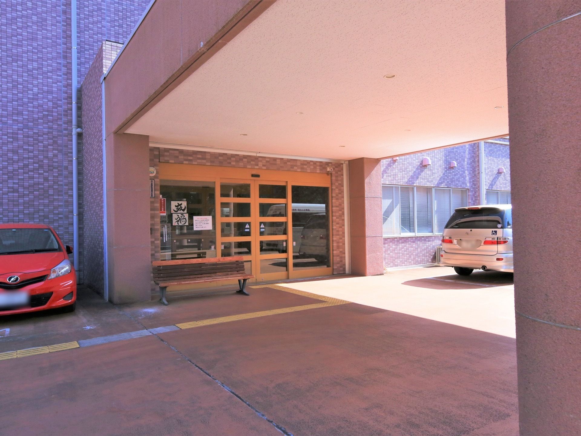 屋根のある車寄せとなっている。自動ドアの玄関横にベンチが置かれている。玄関両脇に車が２台駐車されている。