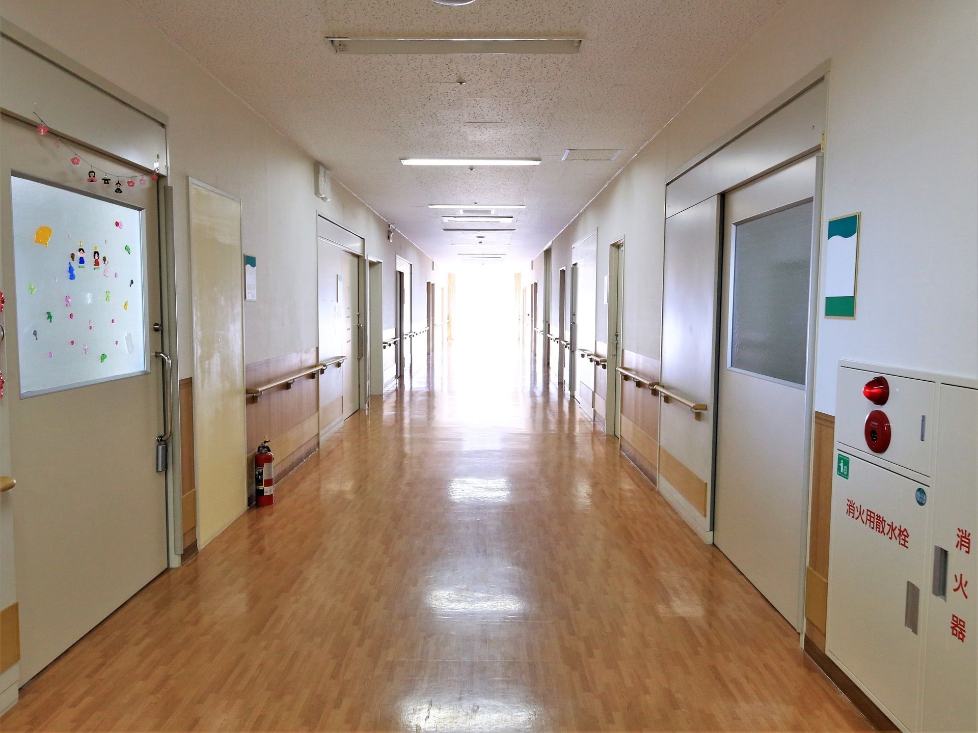 横幅のあるフローリング床の廊下である。両側に引き戸式のドアの部屋があり、壁伝いに手すりが設置されている。