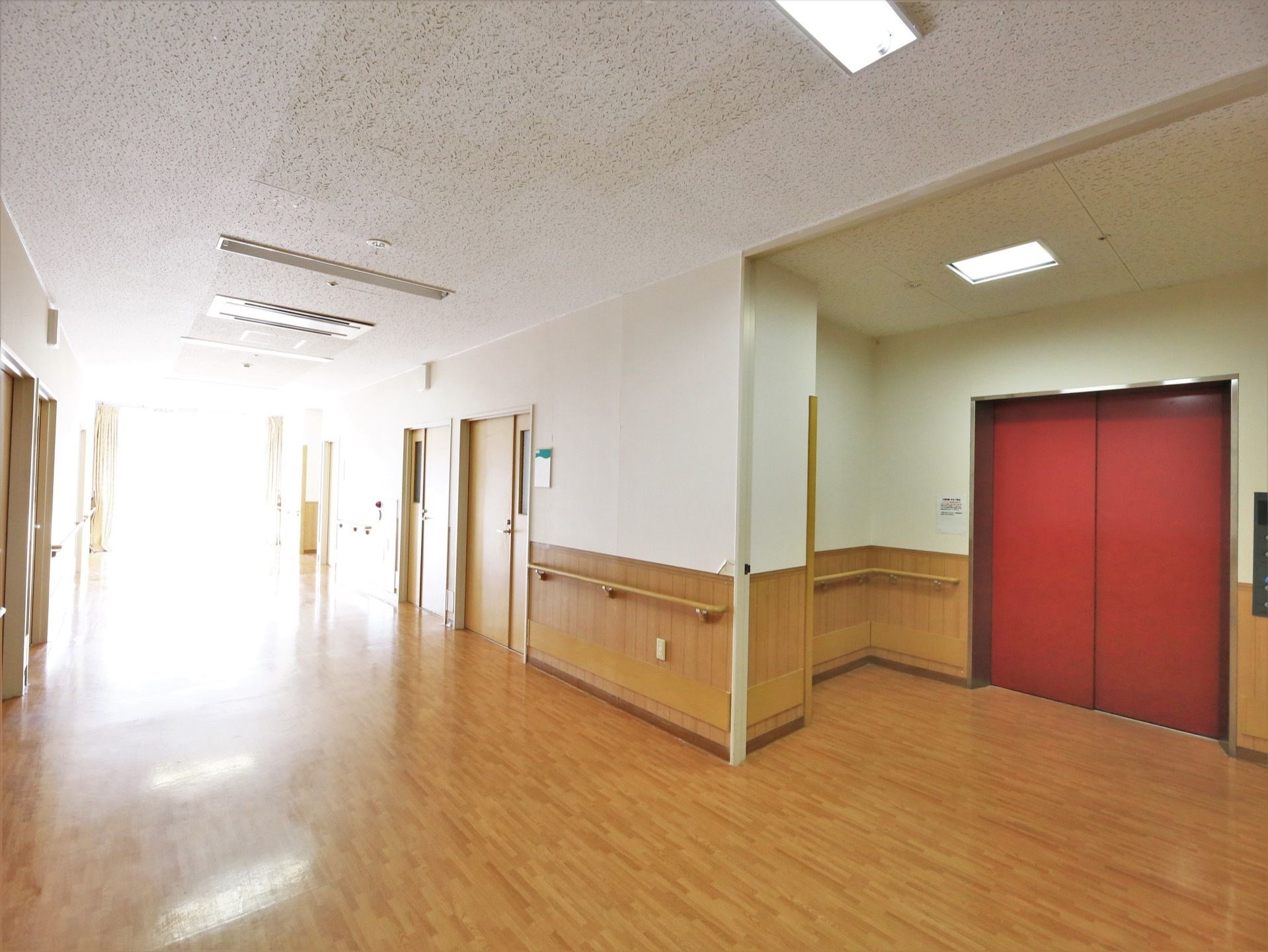 広々とした明るい廊下で、赤い扉のエレベーターが設置されている。廊下には木製の手すりが取りつけられている。