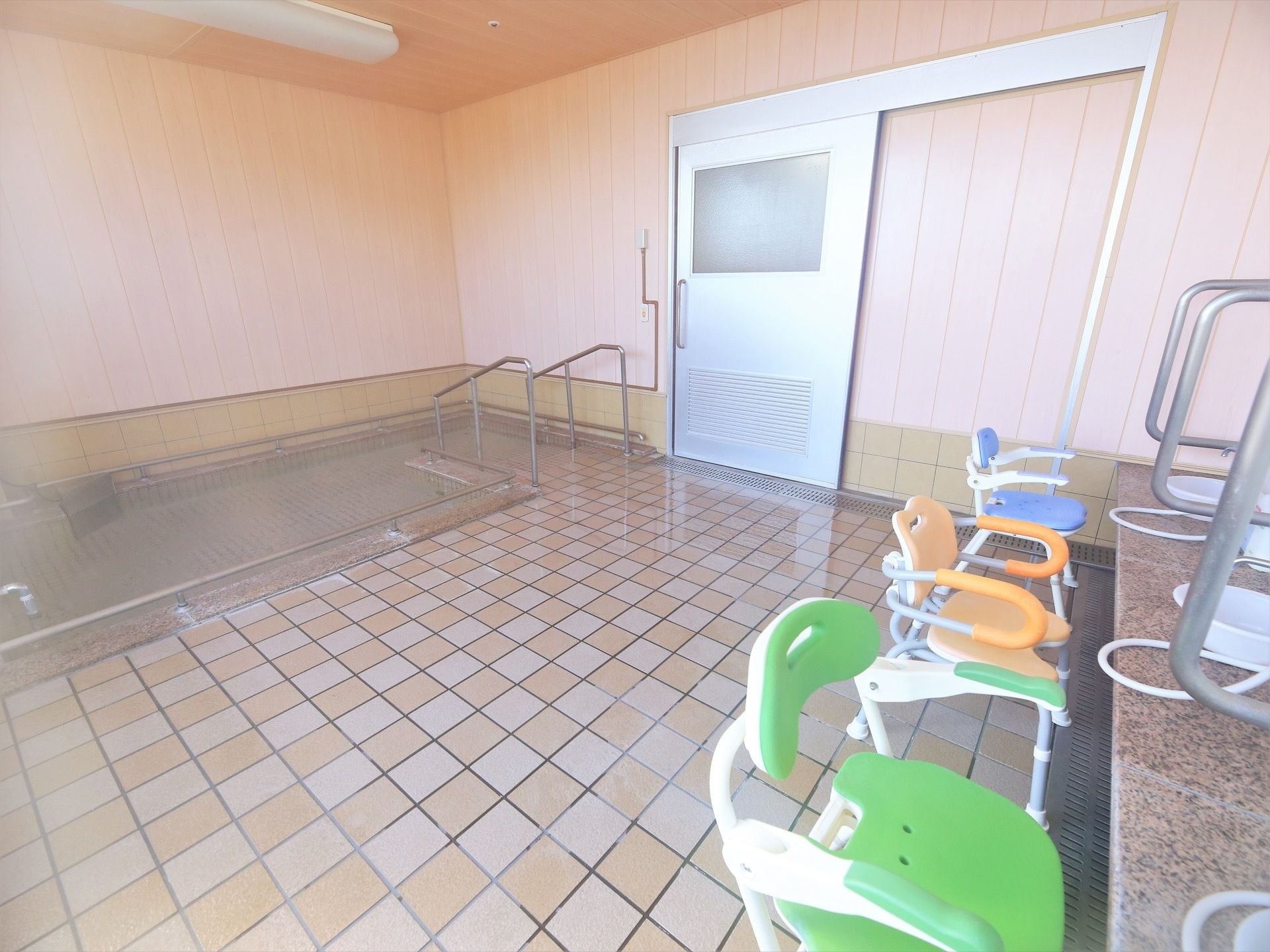 浴室内に湯気が立ち込めている大浴場で、洗い場にはシャワーチェアが置かれている。大浴場の入口はスライド式のドアになっている。