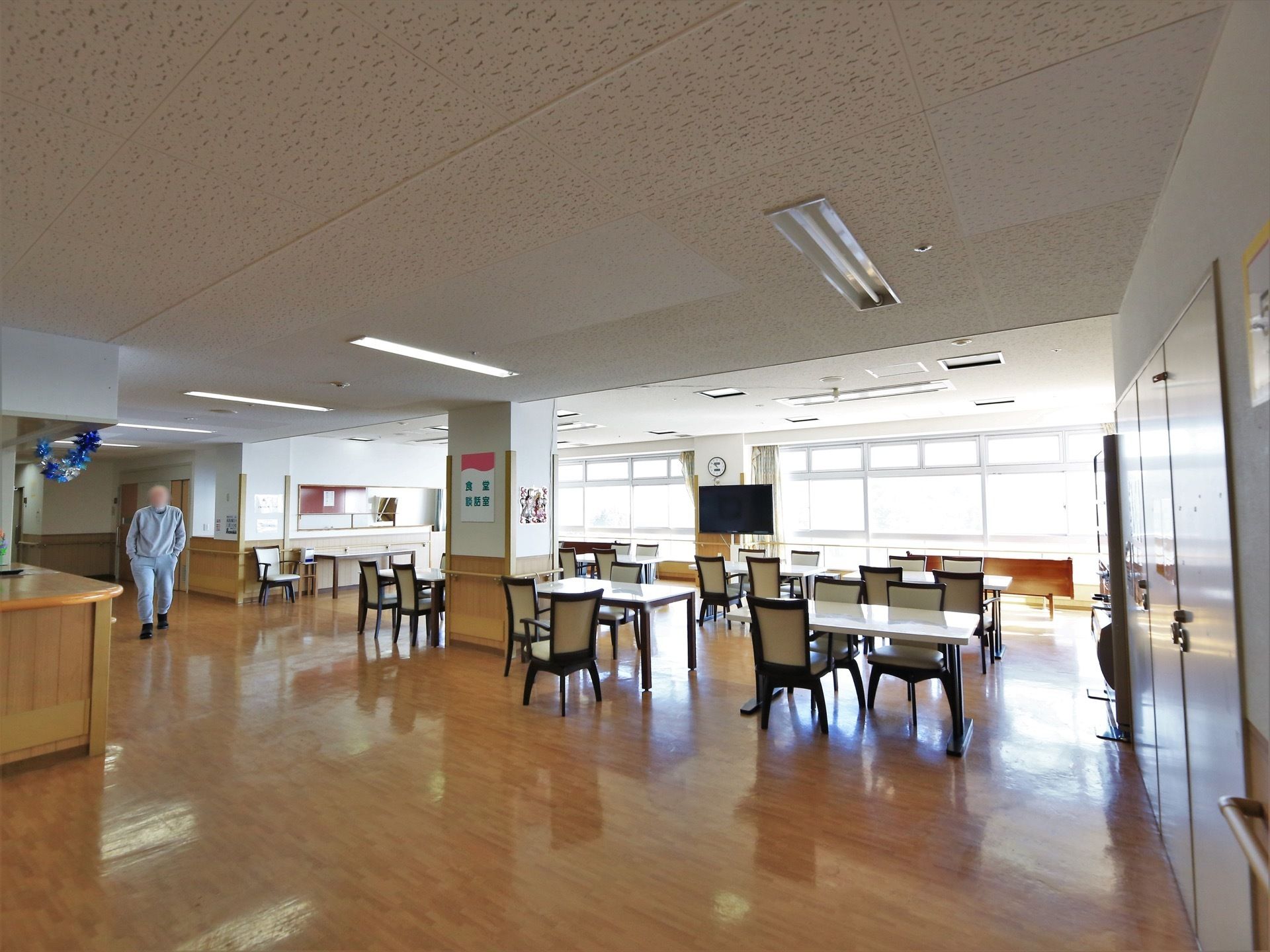 柱に食堂、談話室と書かれた紙が貼られている。窓側にテレビが１台設置されており、後ろの柱に丸型の時計が掛けられている。