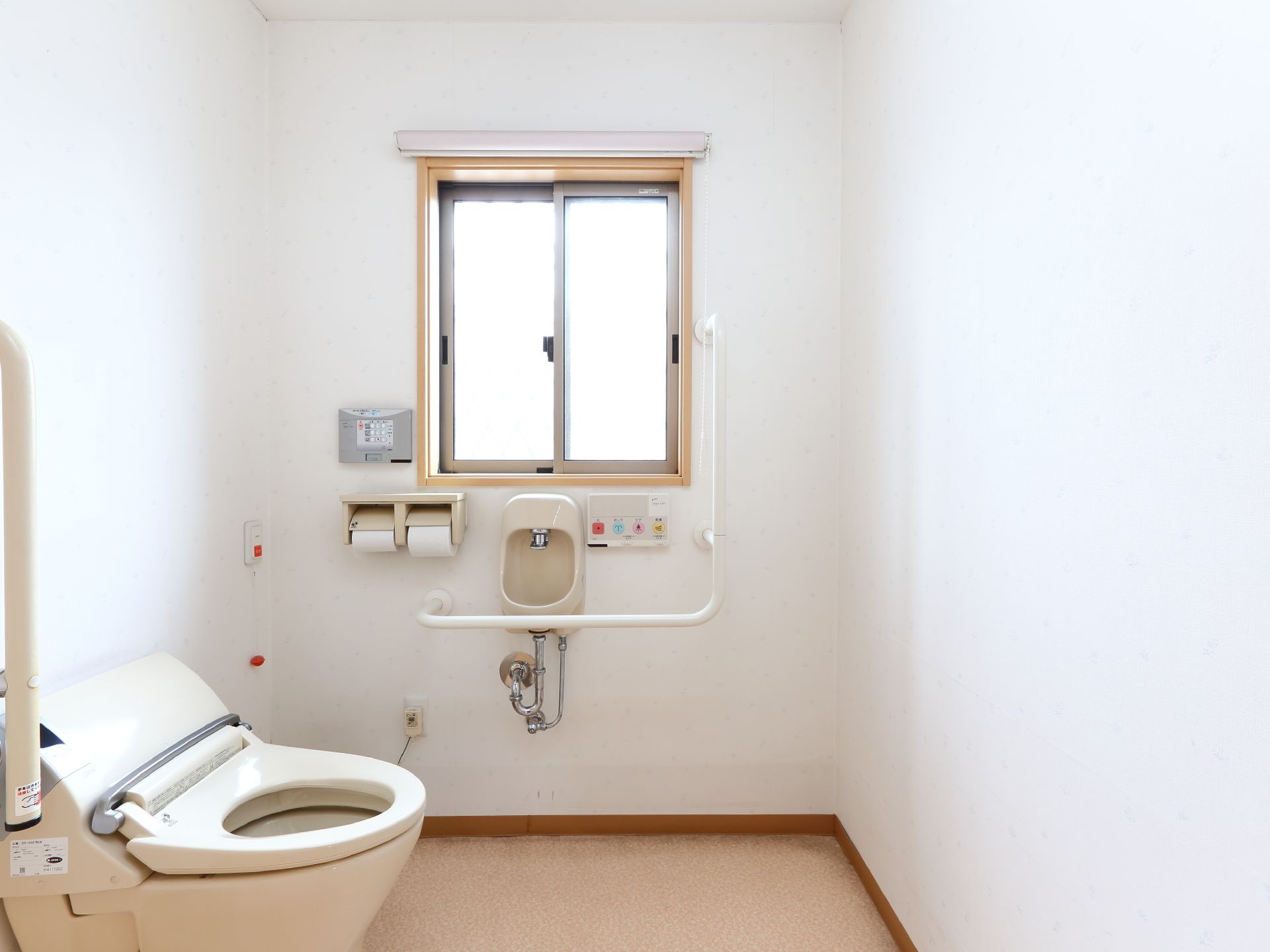 ウオッシュレット式トイレの奥の壁には窓があり日光が差し込んでいる。その下には手すりや水栓に各種スイッチがついている。