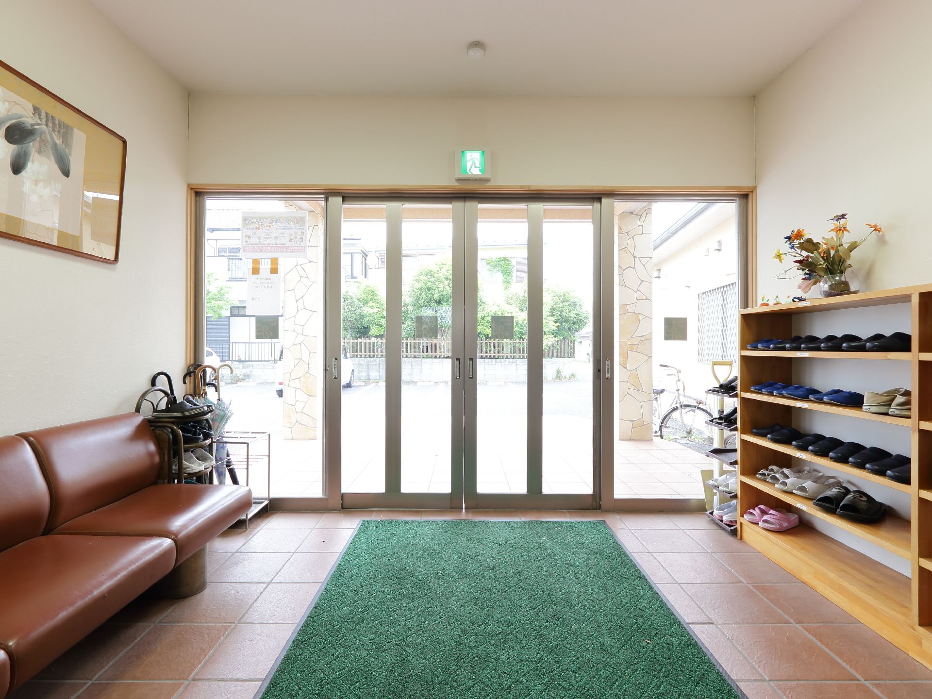 右手には上に花が飾られた下駄箱、右手には茶色のソファと傘立てがある。自動ドアを通りバリアフリーで屋外にでれる。
