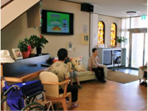 玄関に入ってすぐのところにあるリビング。ソファや椅子が置かれ、入居者様が座ってくつろいでいる。壁にはテレビが設置されている。