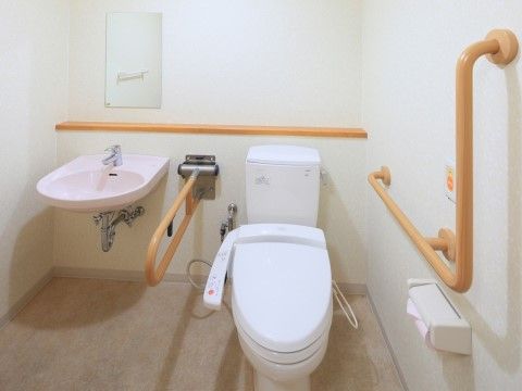 白を基調とした清潔な空間。壁にはトイレ用の手すりが取り付けられている。便器の横には手洗い場を設置している。