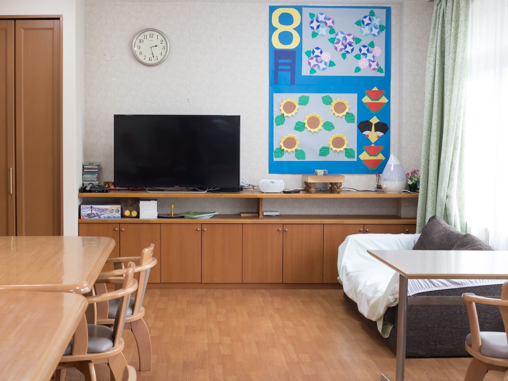 棚の上にテレビが置かれ、壁には時計や折り紙で作成された、かわいらしい掲示物が掛けられている。また、ソファが窓際に置かれている。