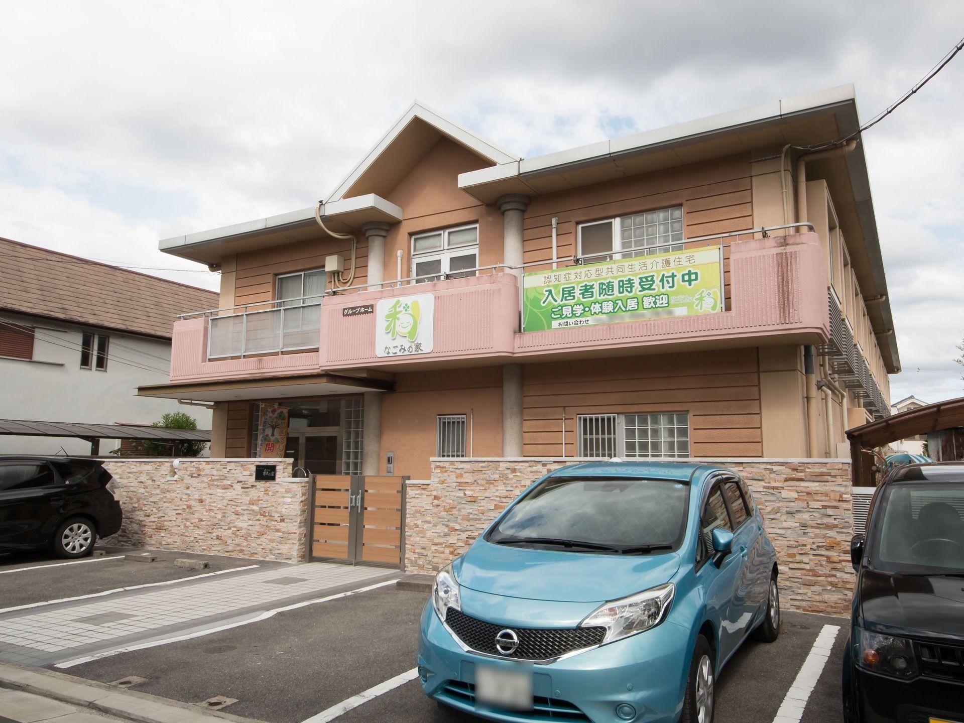 ブロック塀に囲まれた、三角屋根がある２階建ての建物である。塀の前には駐車スペースがあり、車が停まっている。