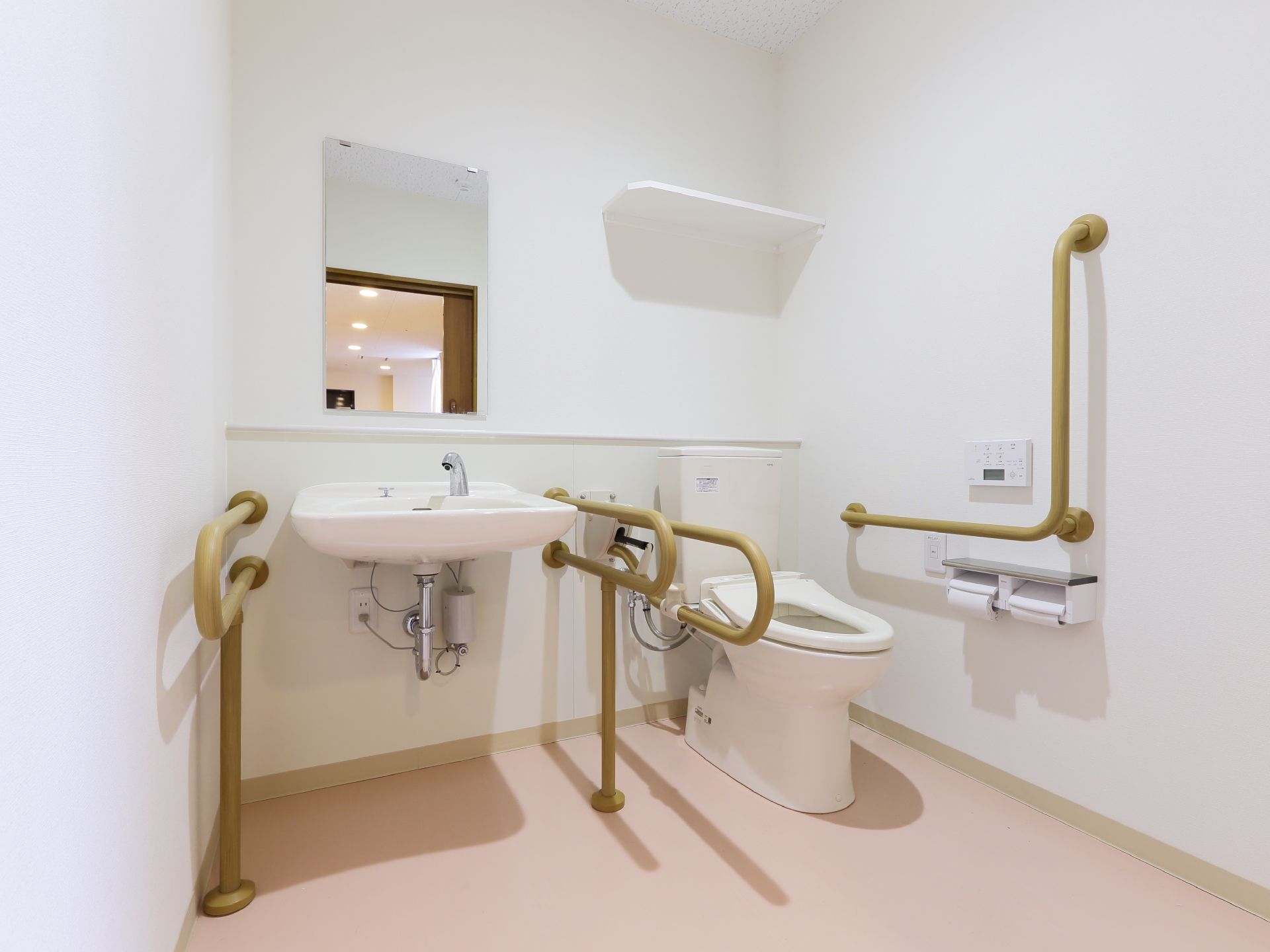 トイレは白壁とピンクの床。ウォシュレットの水洗トイレと洗面台がある。壁と便器と洗面台にはそれぞれ手すりが設置されている。