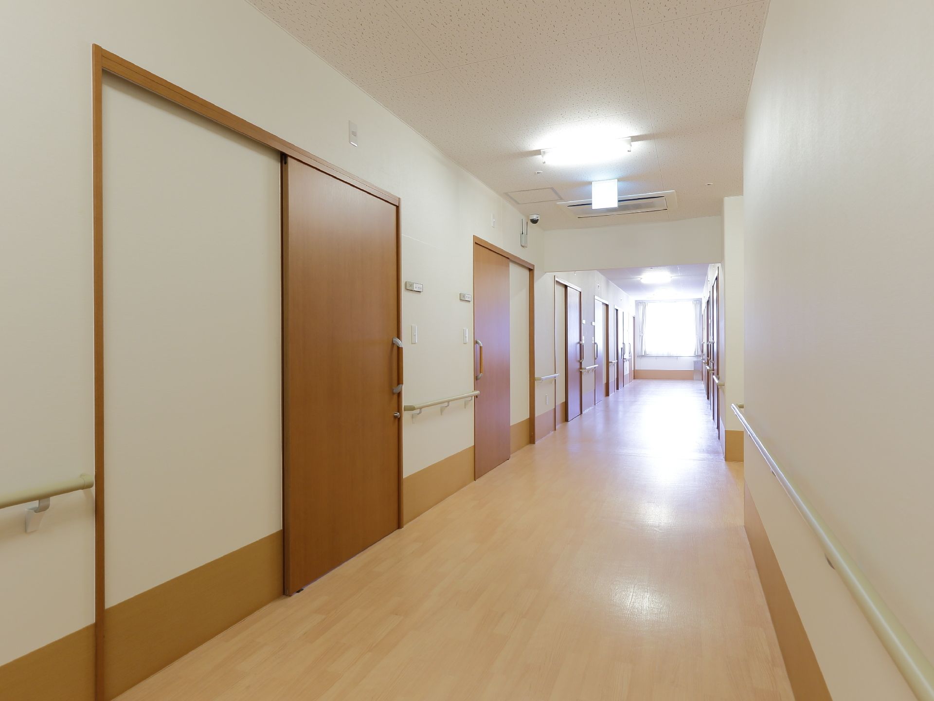 施設の廊下は白壁とフローリングの床。壁には手すりがあり居室の扉が並んでいる。通路は幅広で車椅子でも通行できる。