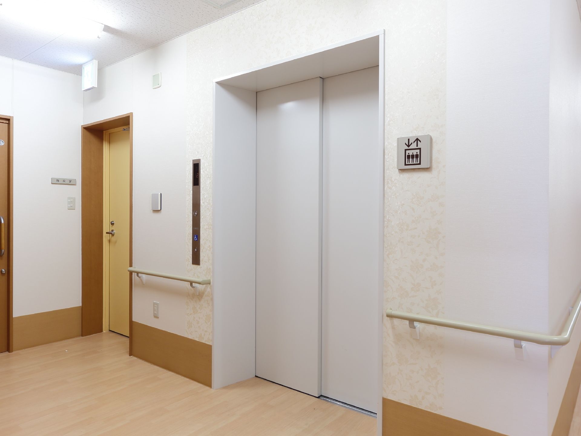 施設には壁の色に合わせた白のエレベーターが設置されている。壁には判りやすくマークとパネルがあり、エレベーターの横には非常階段も見える。
