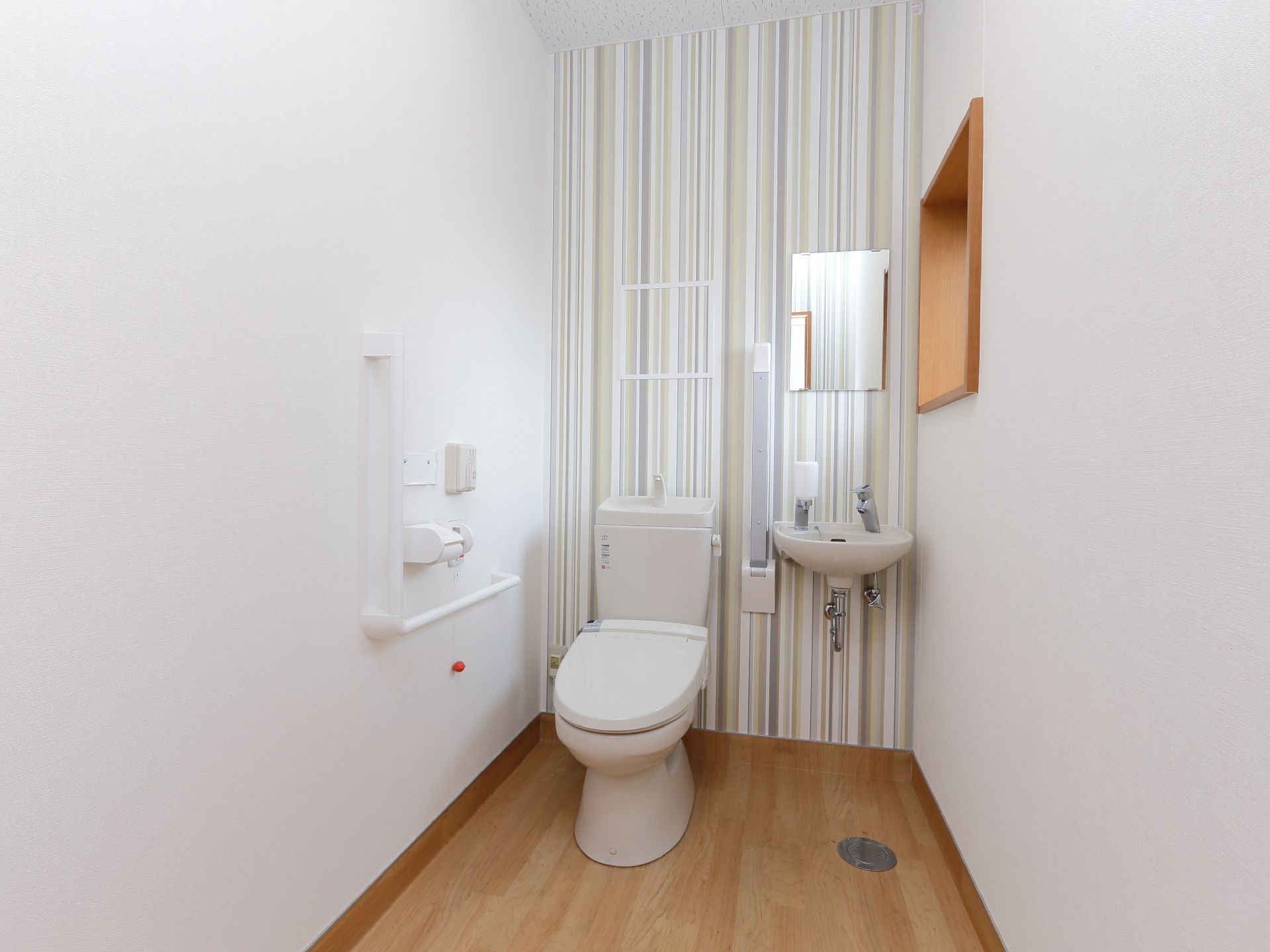 トイレは白壁にフローリング。壁には手すりとスイッチ類があり、便器の横にも可動式の手すりが設置されている。小型の洗面台もある。