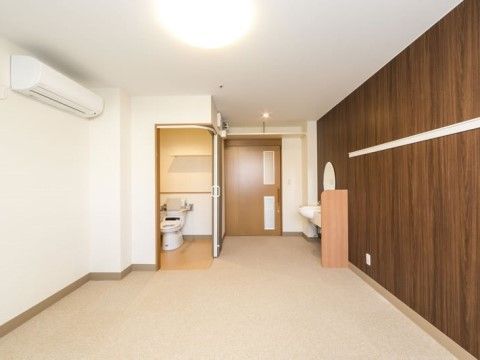 白を基調としたシンプルな部屋の壁の一面が茶色い木目になっている。トイレの入り口の扉が開かれ、洋式便器が見えている。