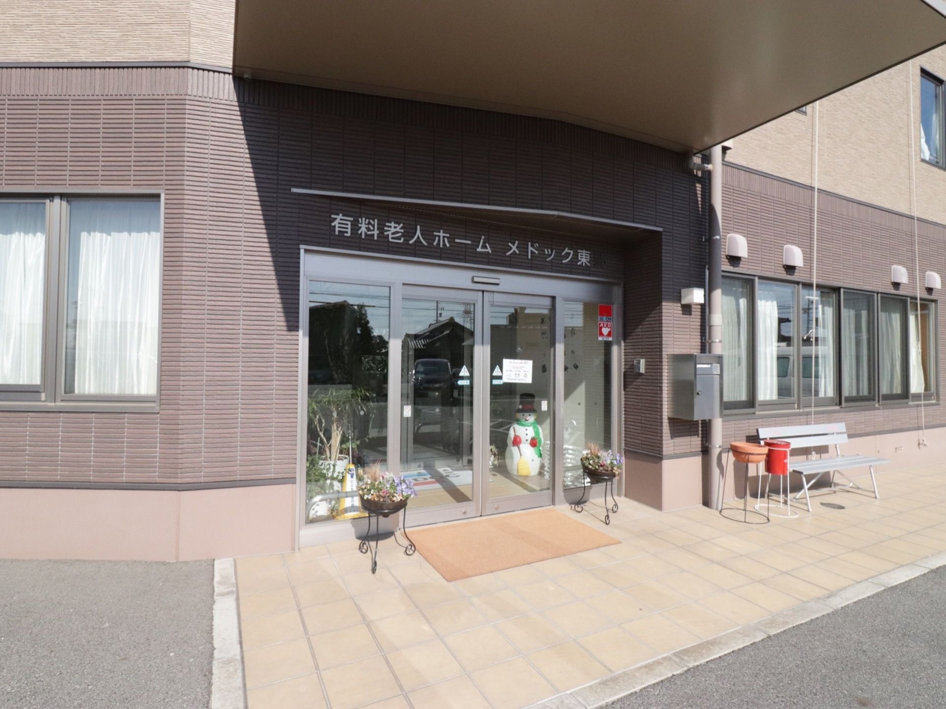 施設の正面玄関。自動ドアはガラス張りのセンサー式スライドダで、右側にはAEDを常備する旨のステッカーが貼られている。