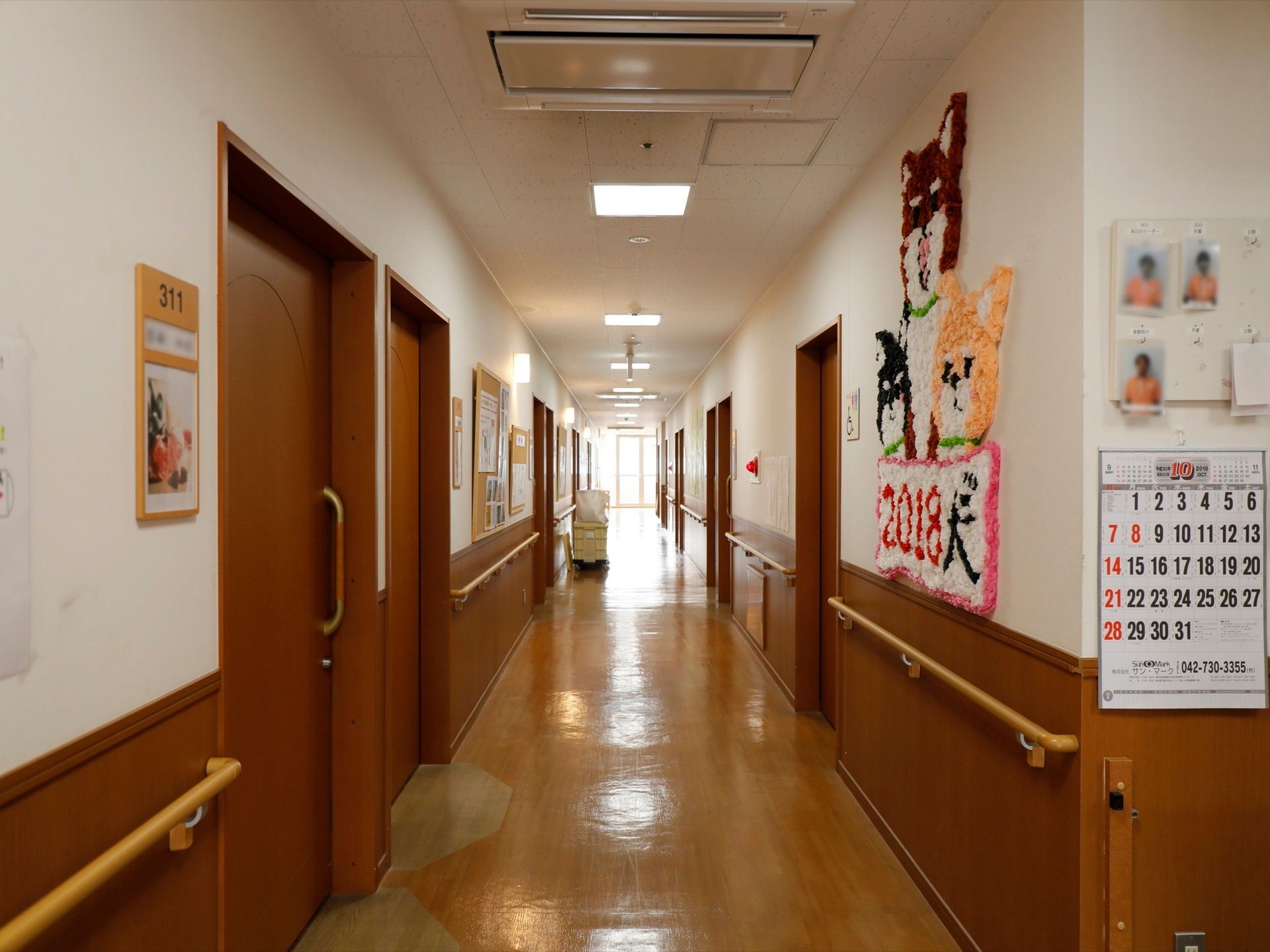 廊下にスタッフの顔写真、カレンダーや干支の図柄が掲示されている。居室入口には居室番号がはっきりと示されている。