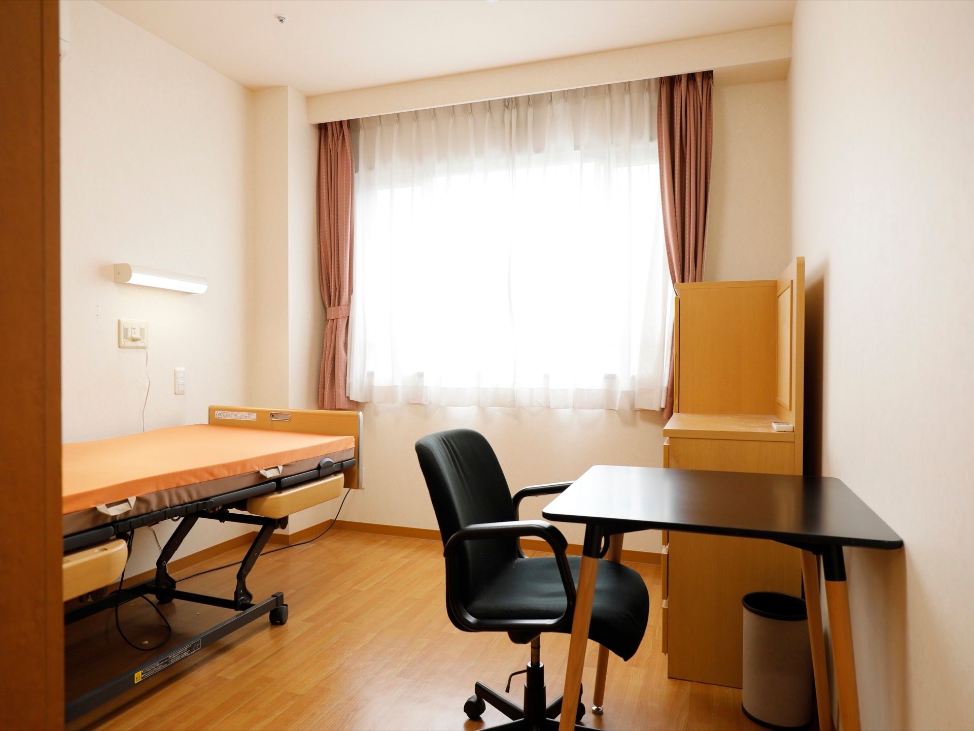 居室に介護ベッドや収納家具、机と椅子が置かれている。床はフローリングになっている。大きな窓にはカーテンがかかっている。