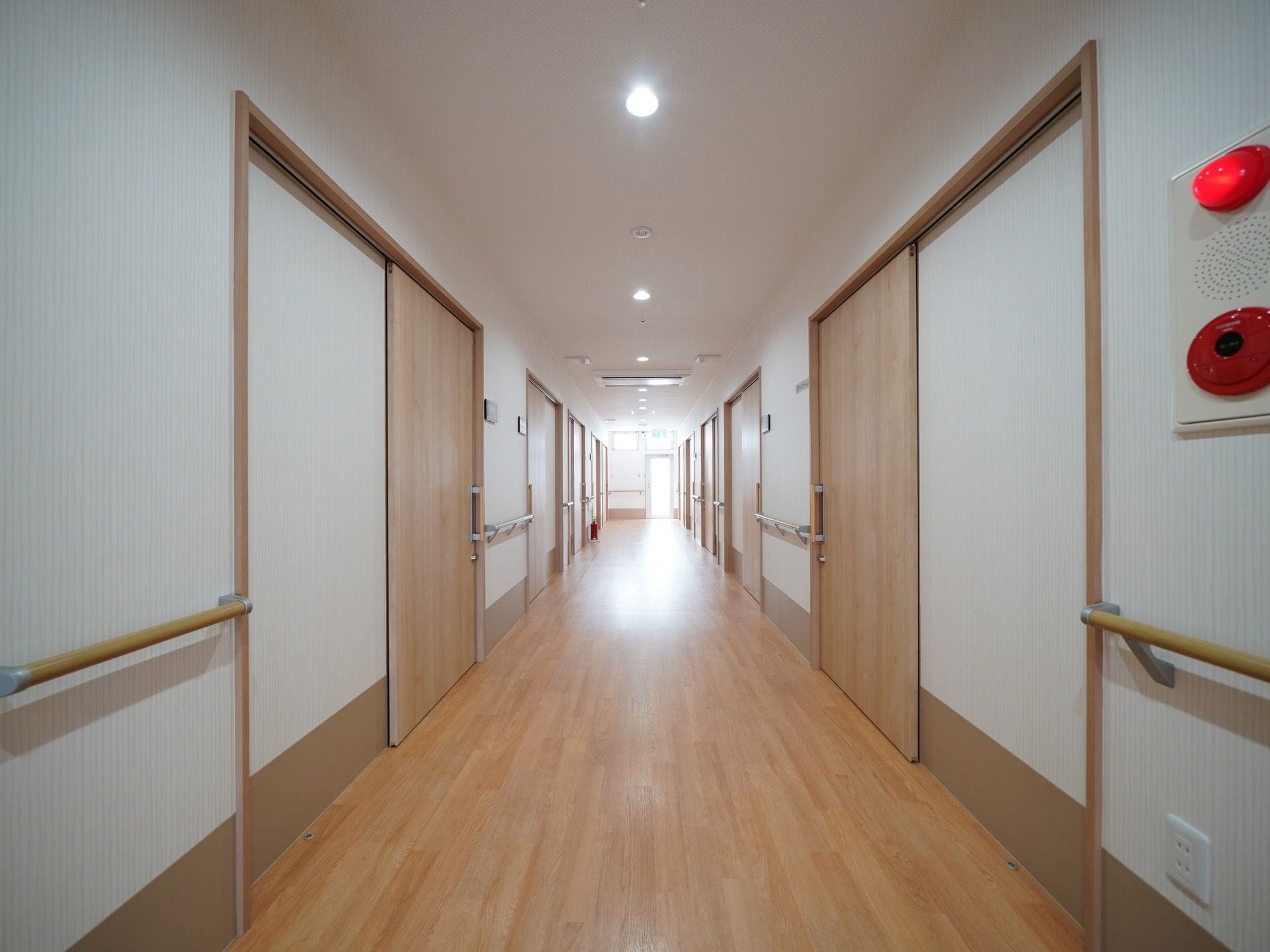 廊下の突き当りにある窓からは外光が差し込んでいる。両側に居室へのドアが並んでいる。壁に非常ベルが設置されている。