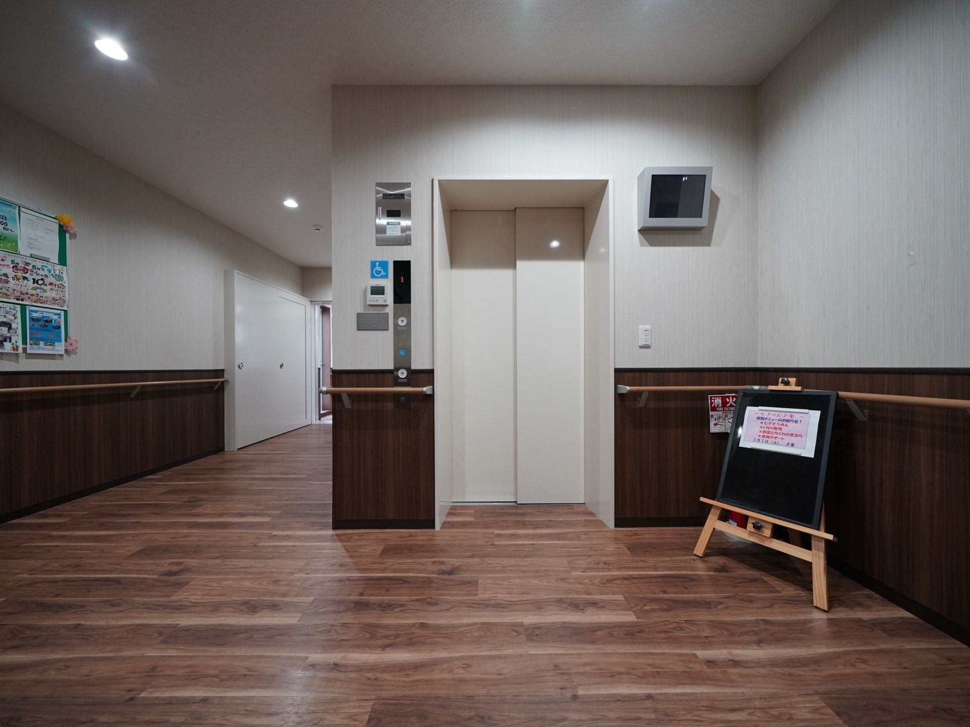 エレベーター前には黒板の立て看板が置かれ、さまざまな情報が貼りだされている。壁に監視カメラのモニターが設置されている。