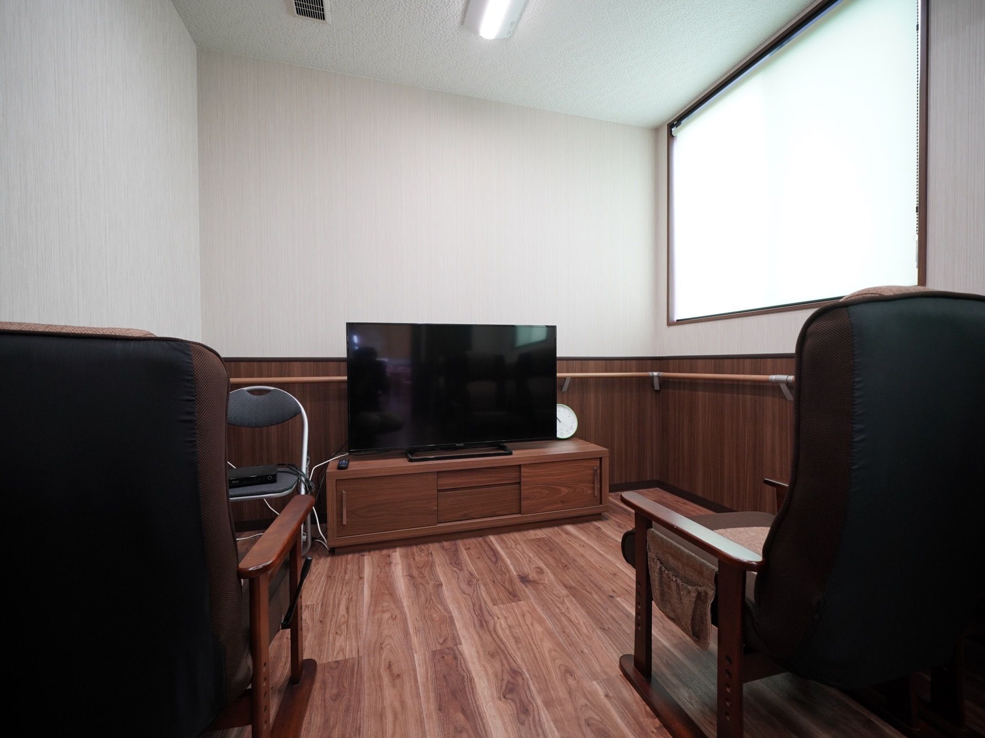 大型液晶テレビが置かれた台の前に、１人掛け用のソファが２脚用意されている。テレビの横にパイプ椅子が１脚ある。
