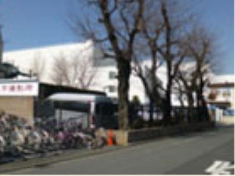 突き当りに白い壁の大きな建物が複数並んでいる。手前には並木があり、駐車スペースがある。看板の前には自転車が多く停まっている。