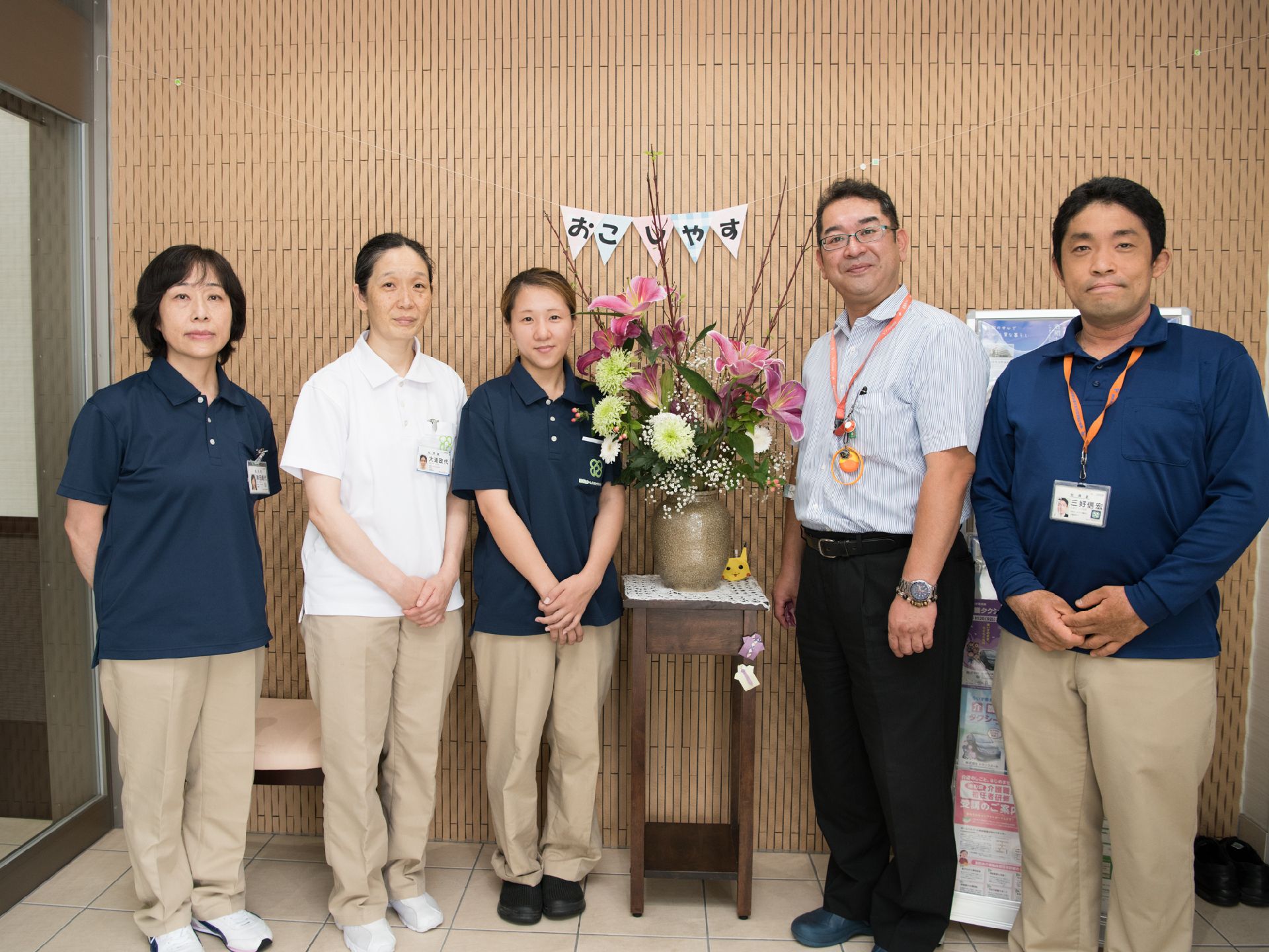 写真には、男性が2名、女性が3名写っている。写真中央には、「おこしやす」と書かれた飾りと、ゆり系の花が花瓶に入れられ、飾られている。