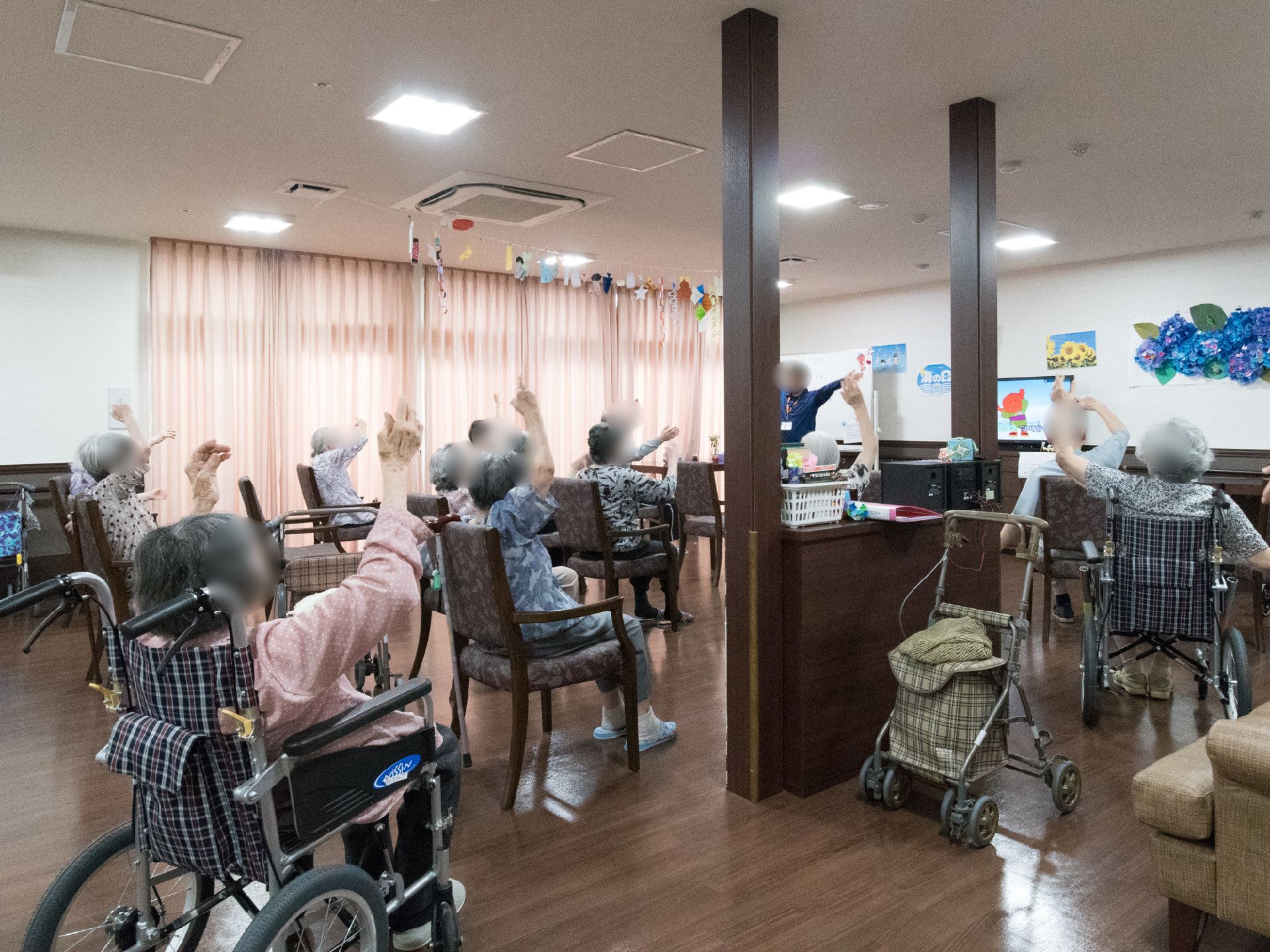 テレビのモニターと職員が前で見本となり、体操を行う。椅子に座ったり、車いすでも行える簡単で楽しみながら体を動かす取り組みをしている。