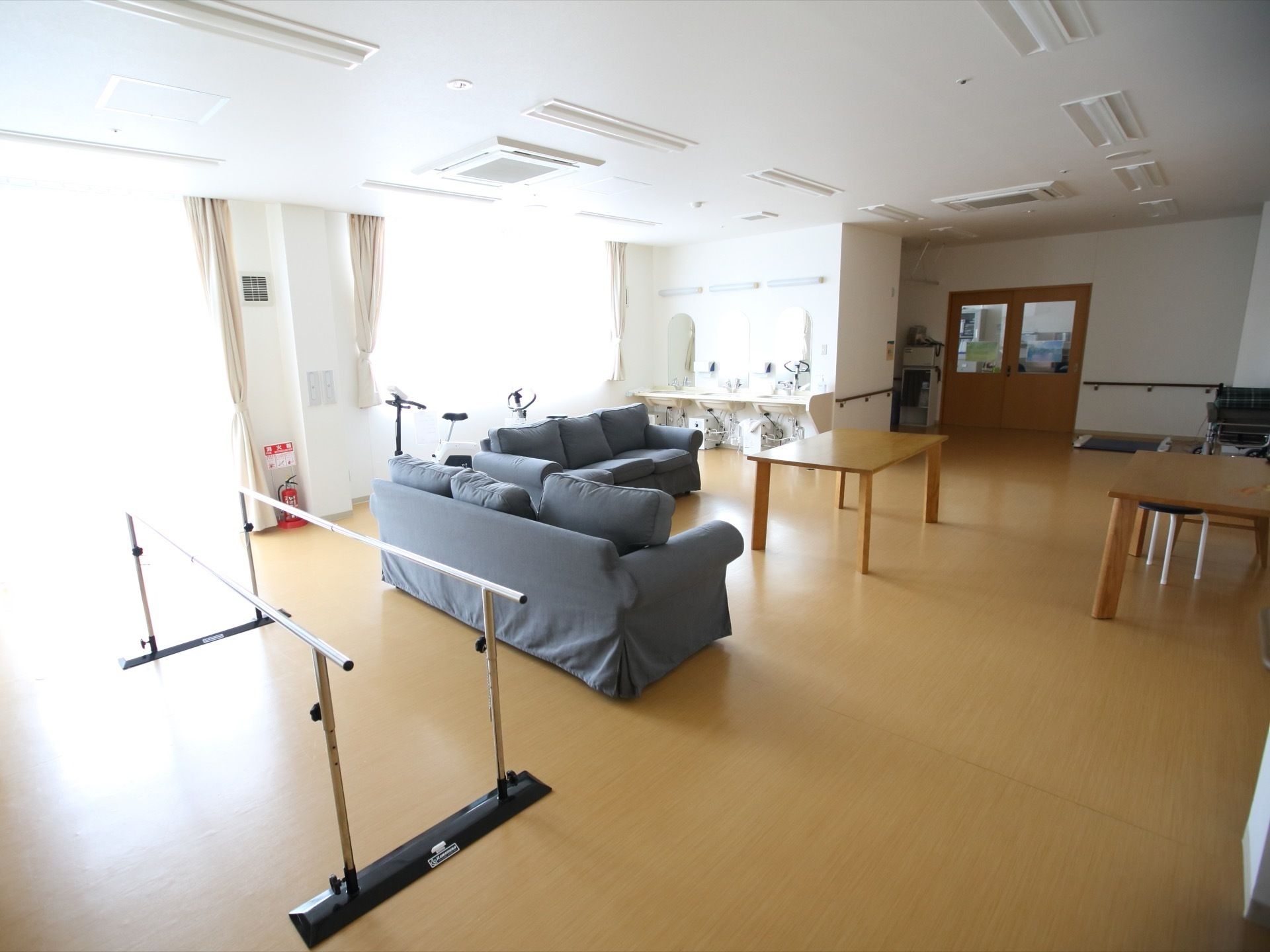 機能訓練室はフローリングの広い空間になっていて、中央にはソファやテーブルが置かれている。まわりには平行棒などのリハビリ機器がある。