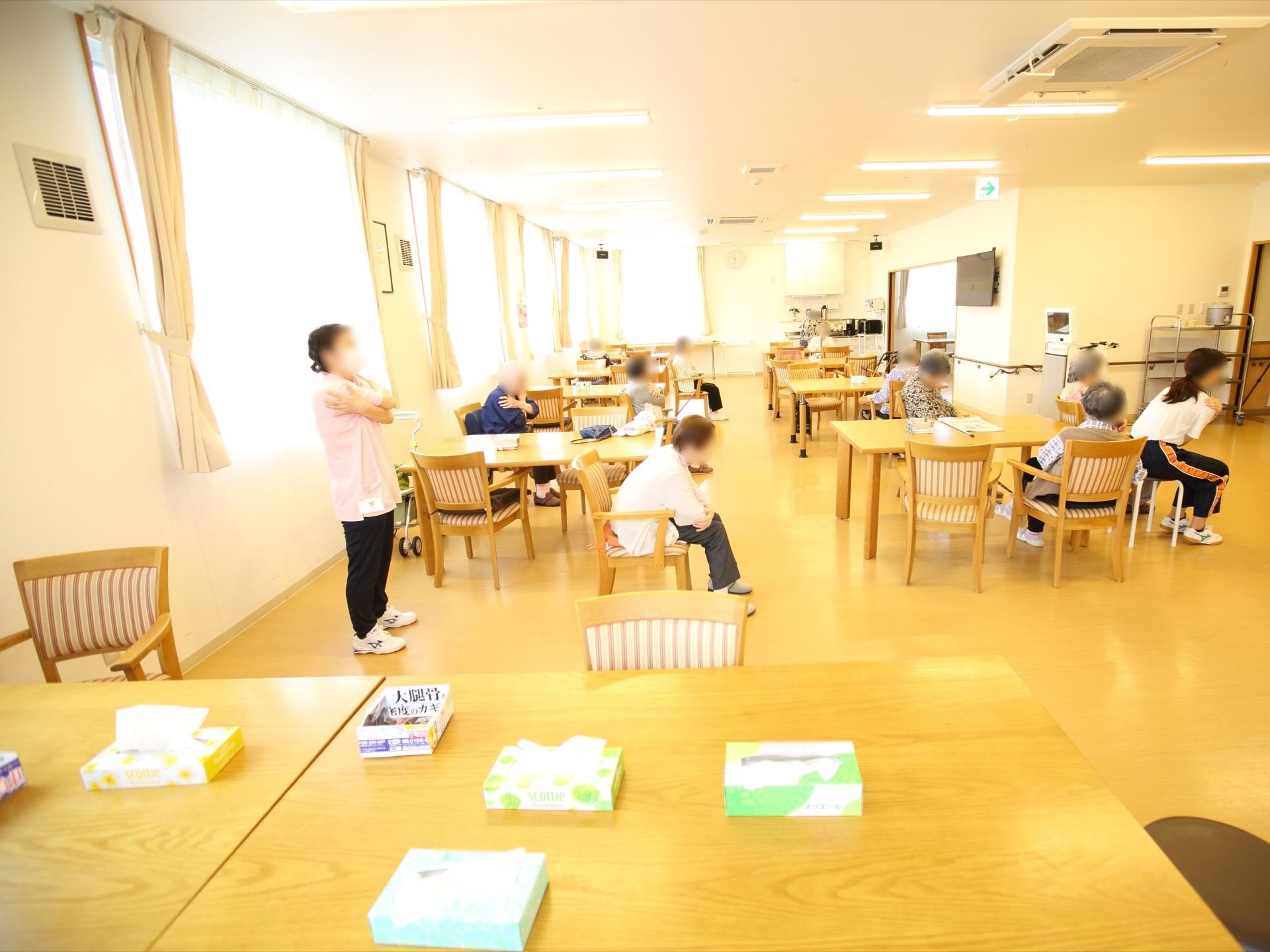 広い空間の食堂にはテーブルと椅子が複数設置されている。入居者様が椅子に座りながらスタッフと一緒に体操をしている。