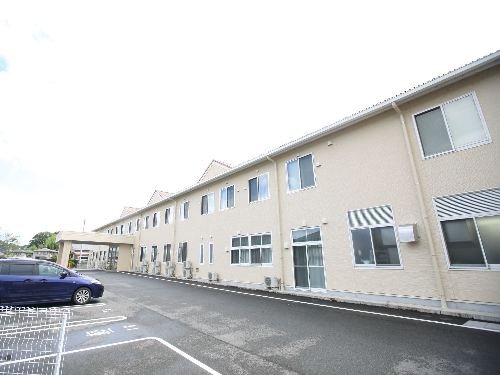 施設は2階建てで、横に長い建物である。建物の前にある駐車場には車が停められており、横には白いフェンスがある。