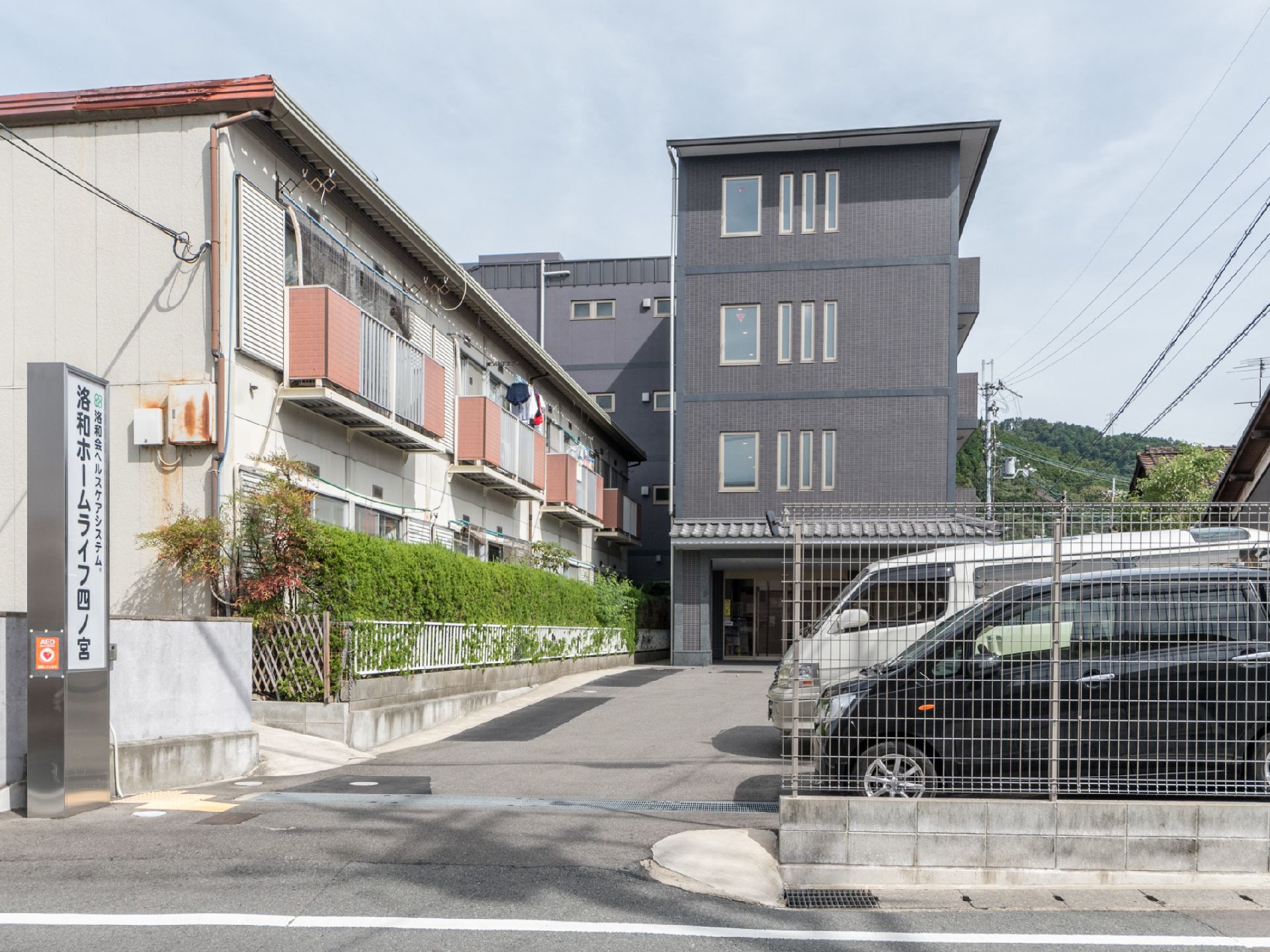道路側の駐車場入り口に縦長の施設の看板が立っている。駐車場の奥に、濃いグレーの4階建ての建物がある。