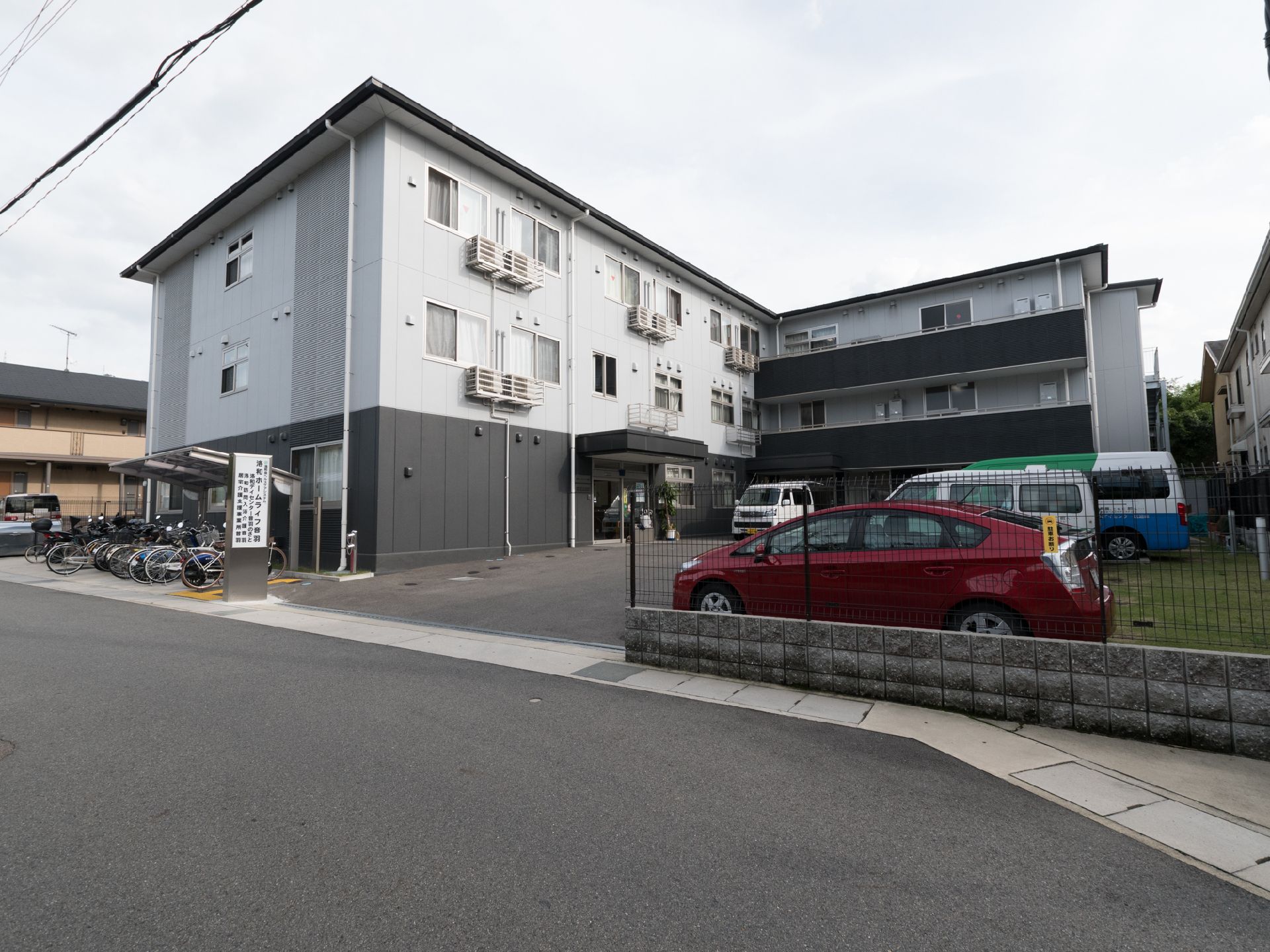 前の道路から段差なく入れる駐車場がある。看板の後ろ側に駐輪場があり、屋根が付いている。建物は3階建てである。