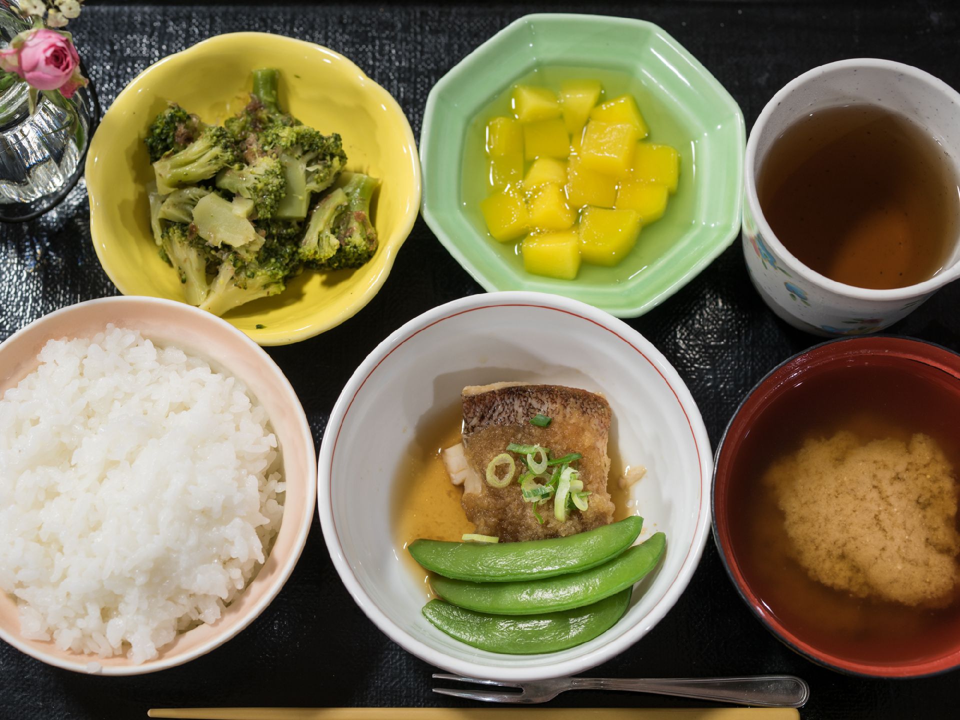 ご飯とお味噌汁に、おかずが二品、デザートの果物がついたメニューである。お茶とともに黒いトレーに載っている。