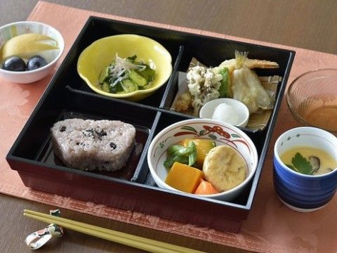 お弁当箱風の器に盛りつけられた食事。天ぷらや根菜の煮物、きゅうりの和え物のほか、茶わん蒸しやフルーツもついている。