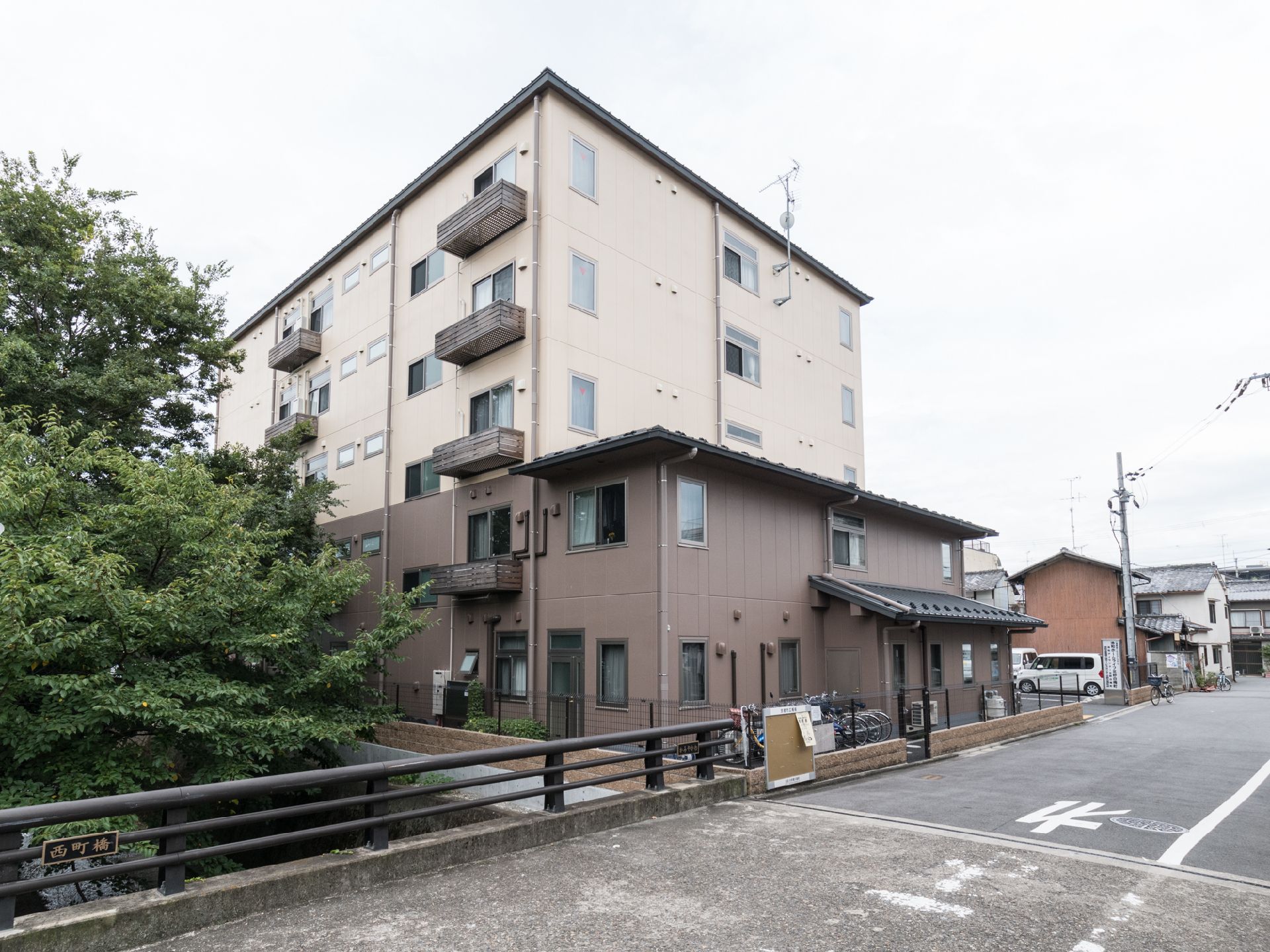 施設の裏には小さな川がある。各階にベランダのついた窓がある。二階までは茶色の外壁で、三階以上はクリーム色である。