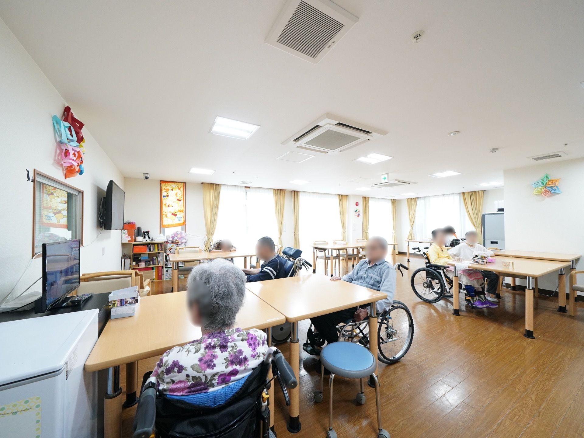 大きな窓のある明るい部屋に、複数のテーブルが並んでいる。車椅子をご利用の入居者様が、数名でテレビを観たり、お茶を飲んだりしている。
